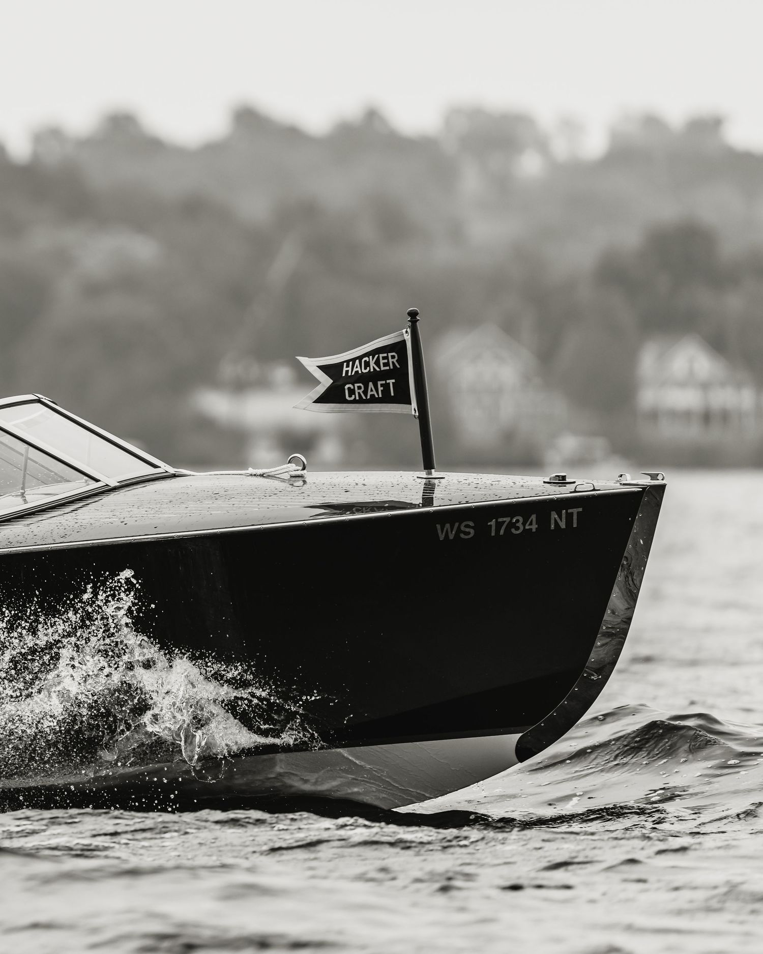 Black and white photo of a Hacker-Craft wooden speedboat with flag, registration WS 1734 NT, cutting through water.