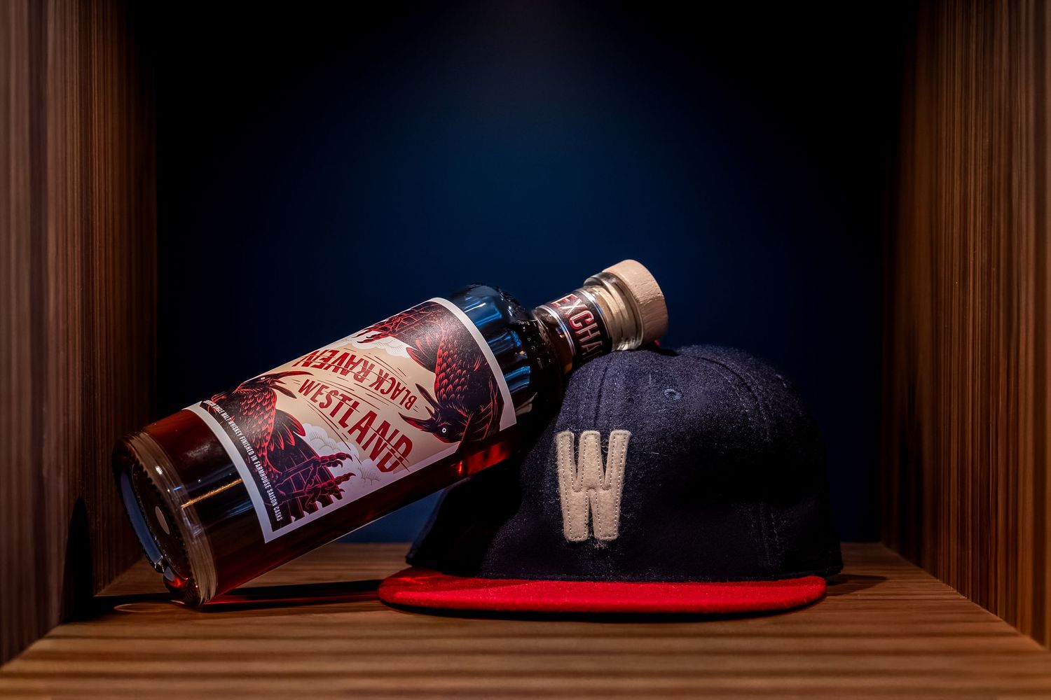 Vintage whiskey bottle and navy W baseball cap displayed on wooden shelf in dim lighting.