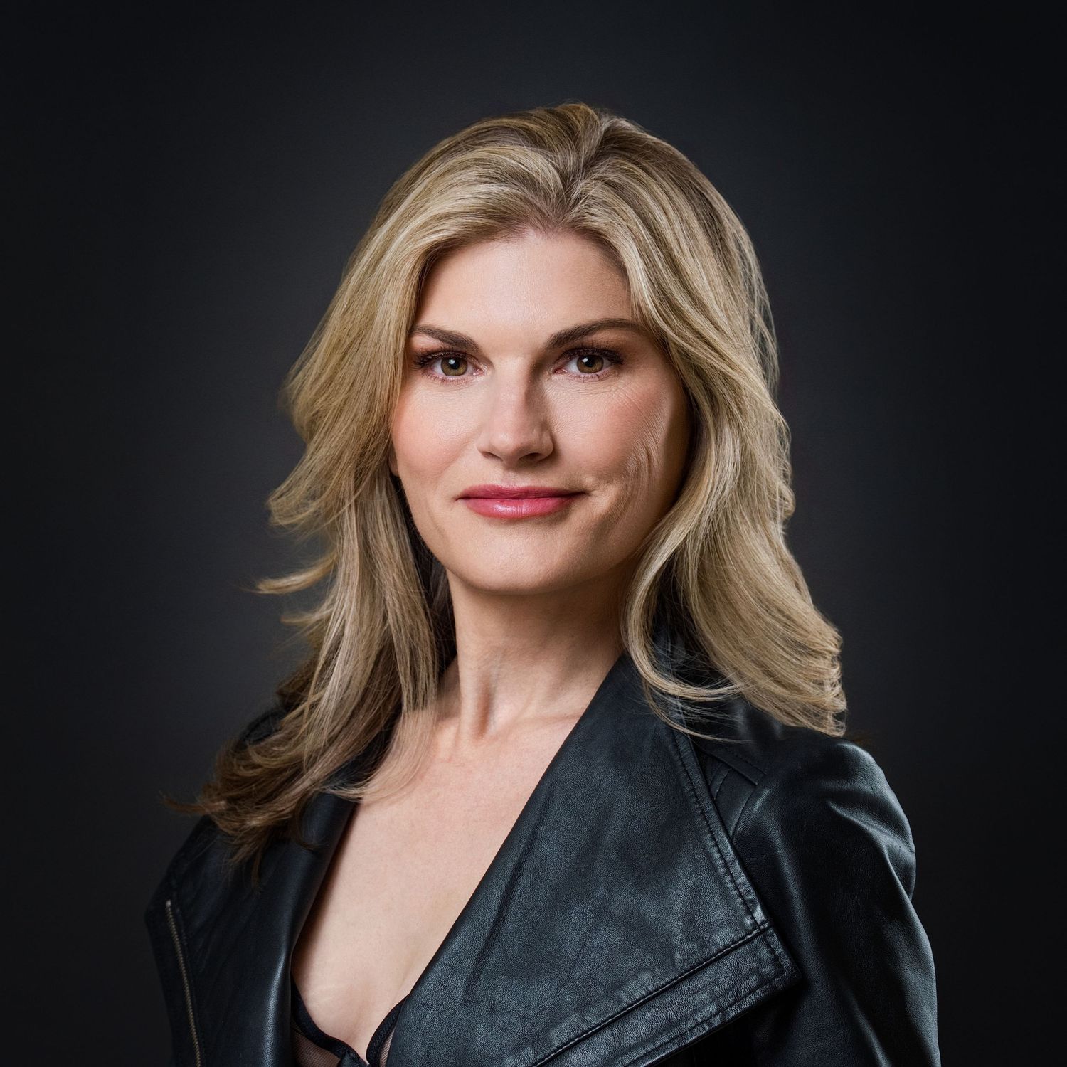 Professional headshot of a confident blonde woman in a black leather jacket against a dark studio background.