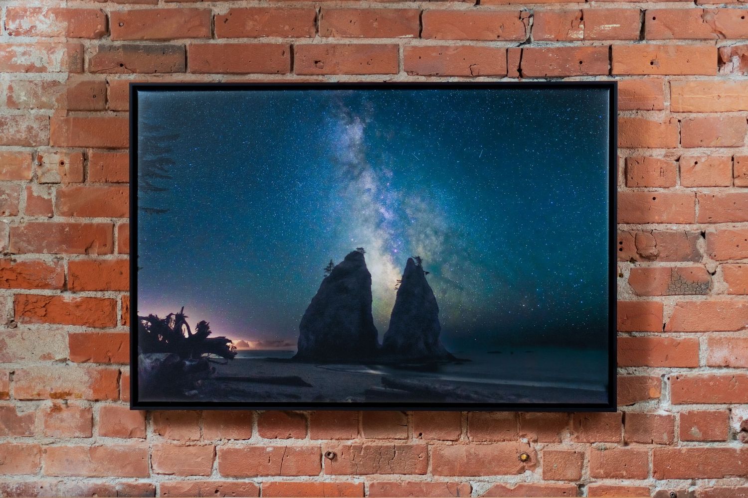 Framed photo of sea stacks under a stunning Milky Way night sky, displayed on a rustic brick wall.