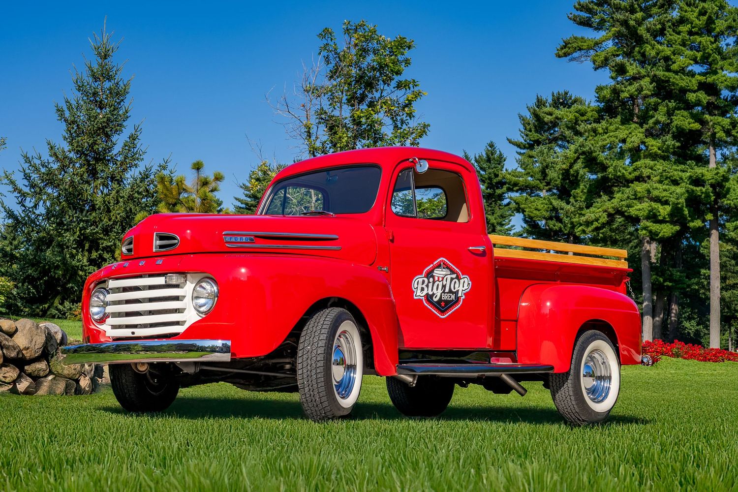 Restored vintage red Ford pickup truck with white wall tires parked on lush green grass under a clear blue sky.