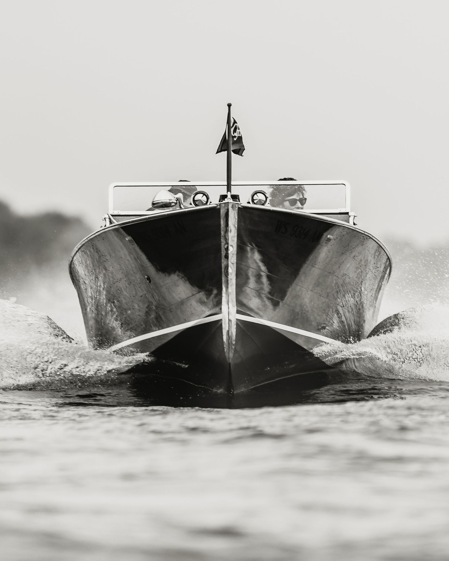 Black and white photo of a classic speedboat racing head-on through choppy water with a flag at the bow.