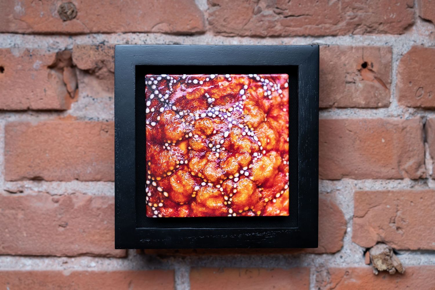 Framed artwork of vibrant orange shrimp dish with white pearls, displayed on a rustic red brick wall.