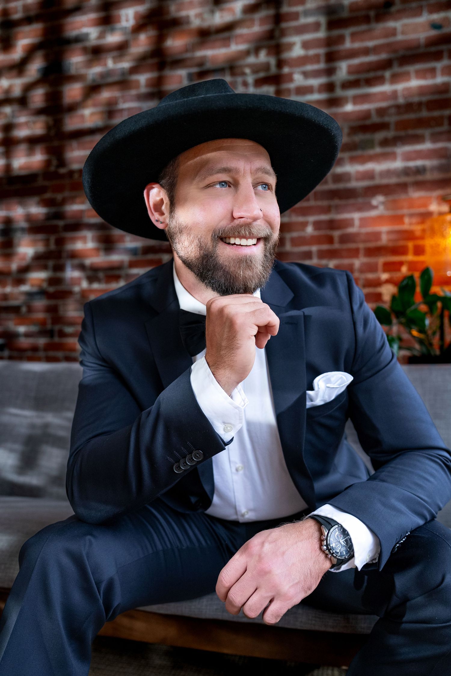 Smiling bearded man in dark suit and black hat posing in brick-walled interior.