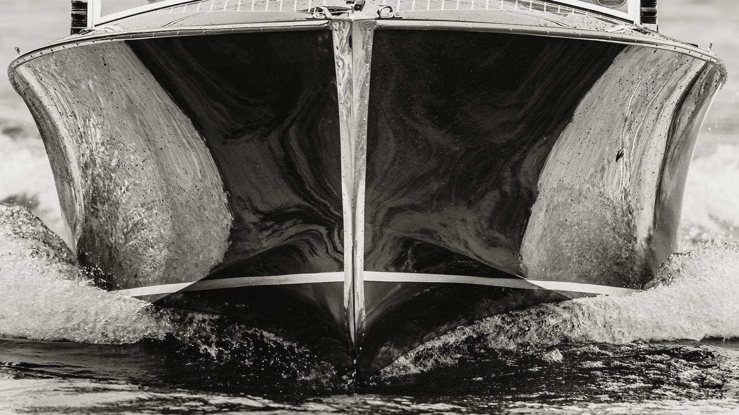 Black and white close-up of a classic wooden speedboat bow cutting through water at high speed.