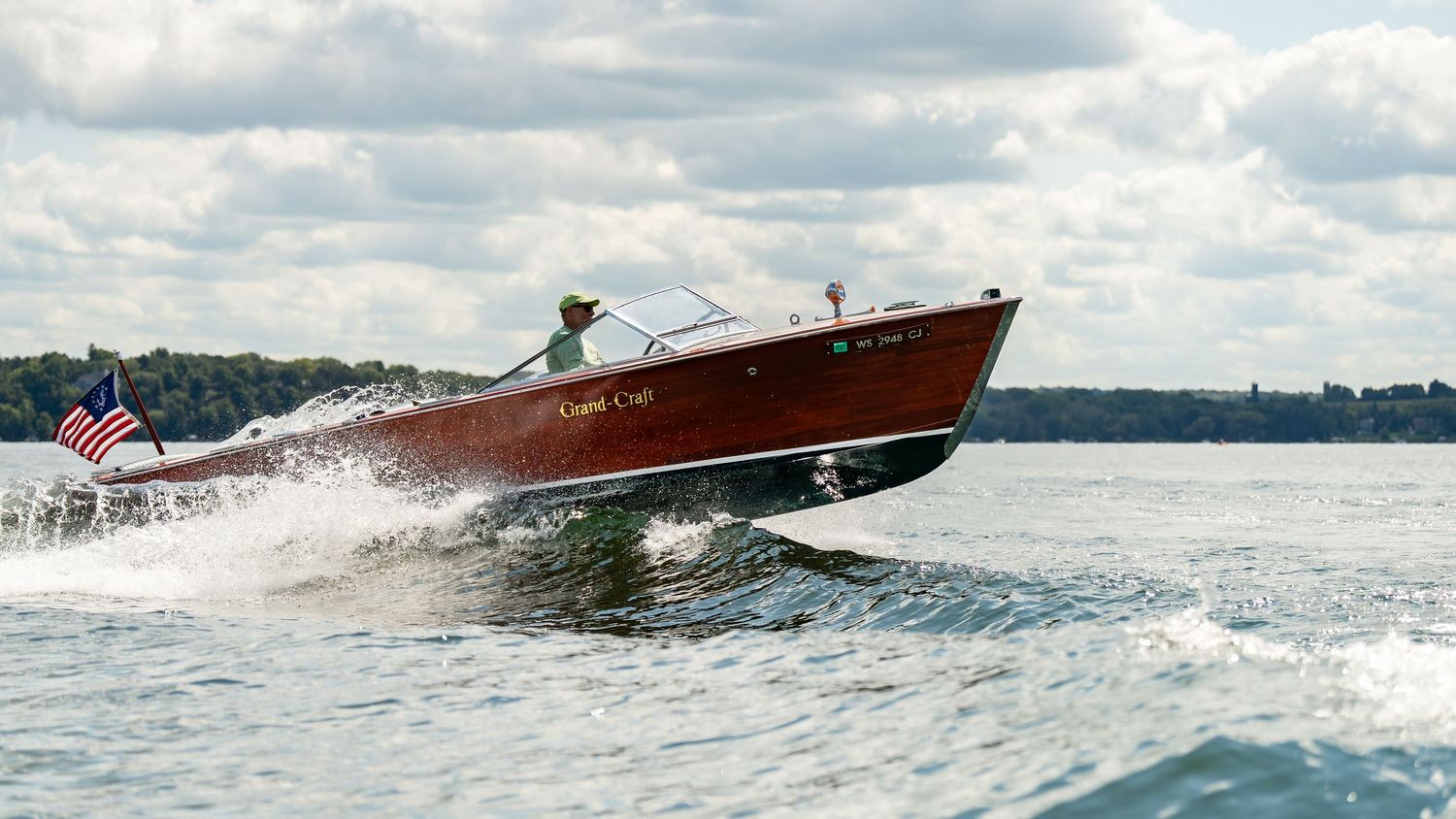 A classic wooden speedboat with an American flag races across calm blue waters under a partly cloudy sky.