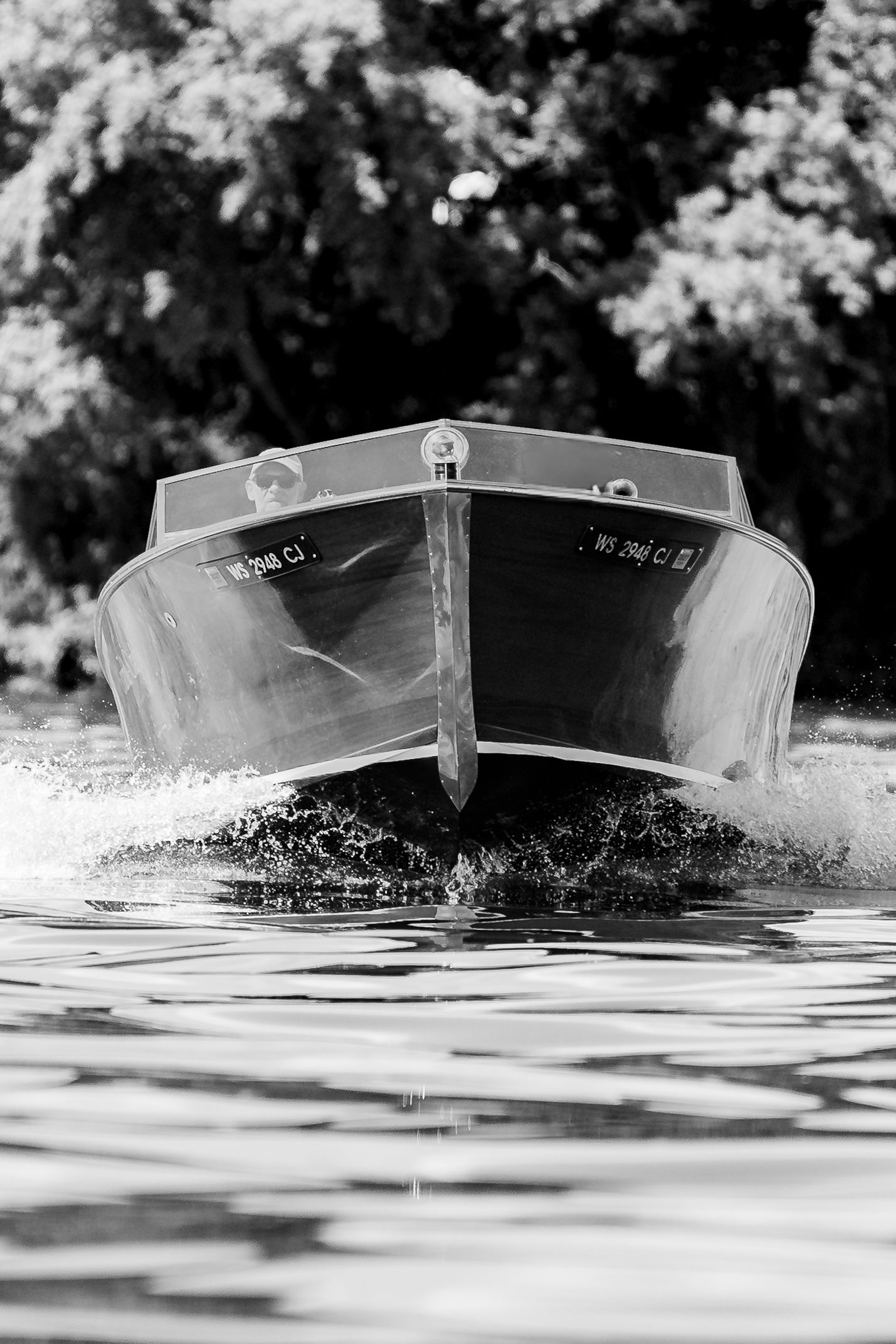Black and white front view of a classic motorboat cutting through water with trees in background.
