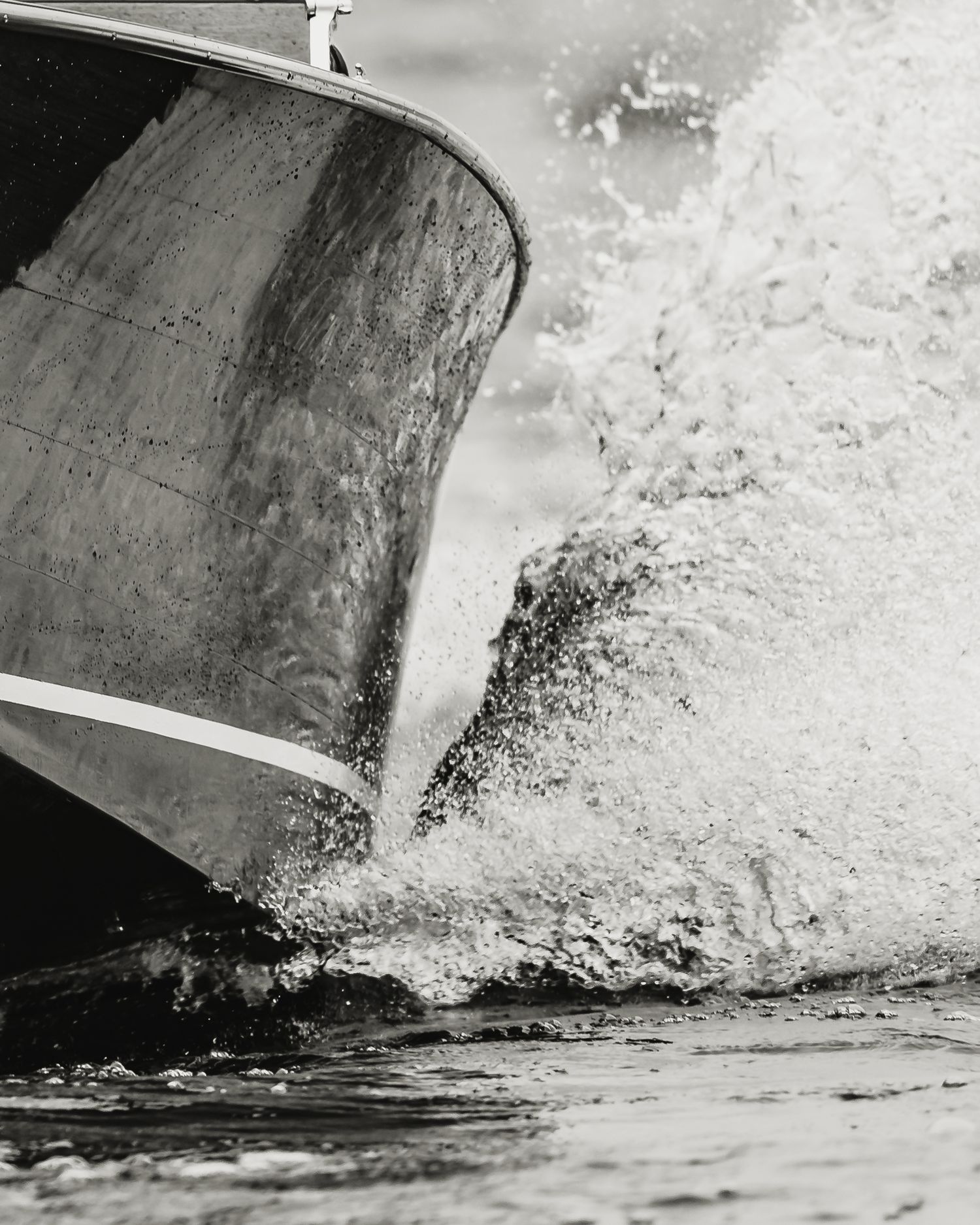 Black and white photo of a boat bow cutting through waves, creating dramatic white water splashes.