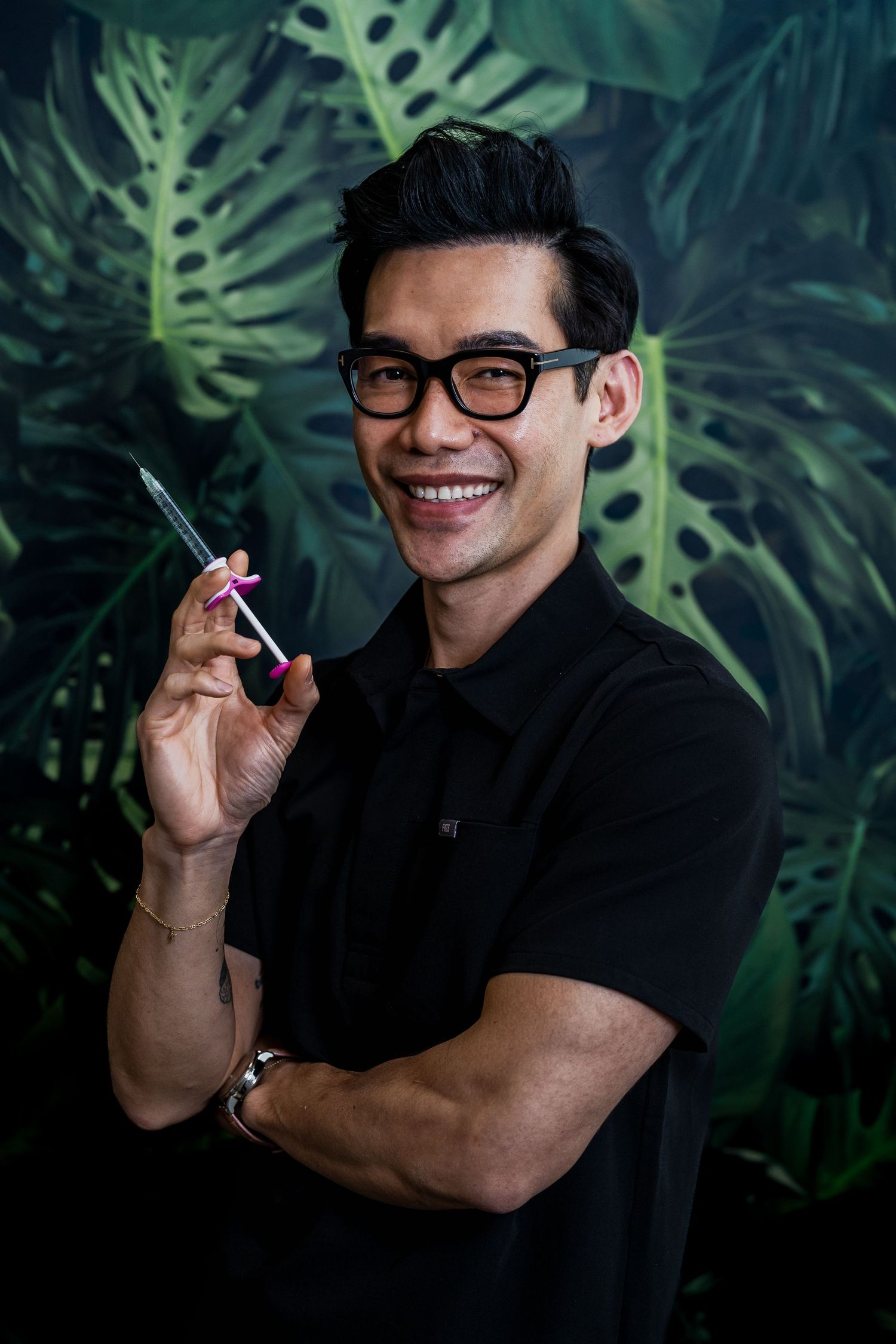 Smiling Asian man in black shirt and glasses holding a pen against tropical monstera leaf background.