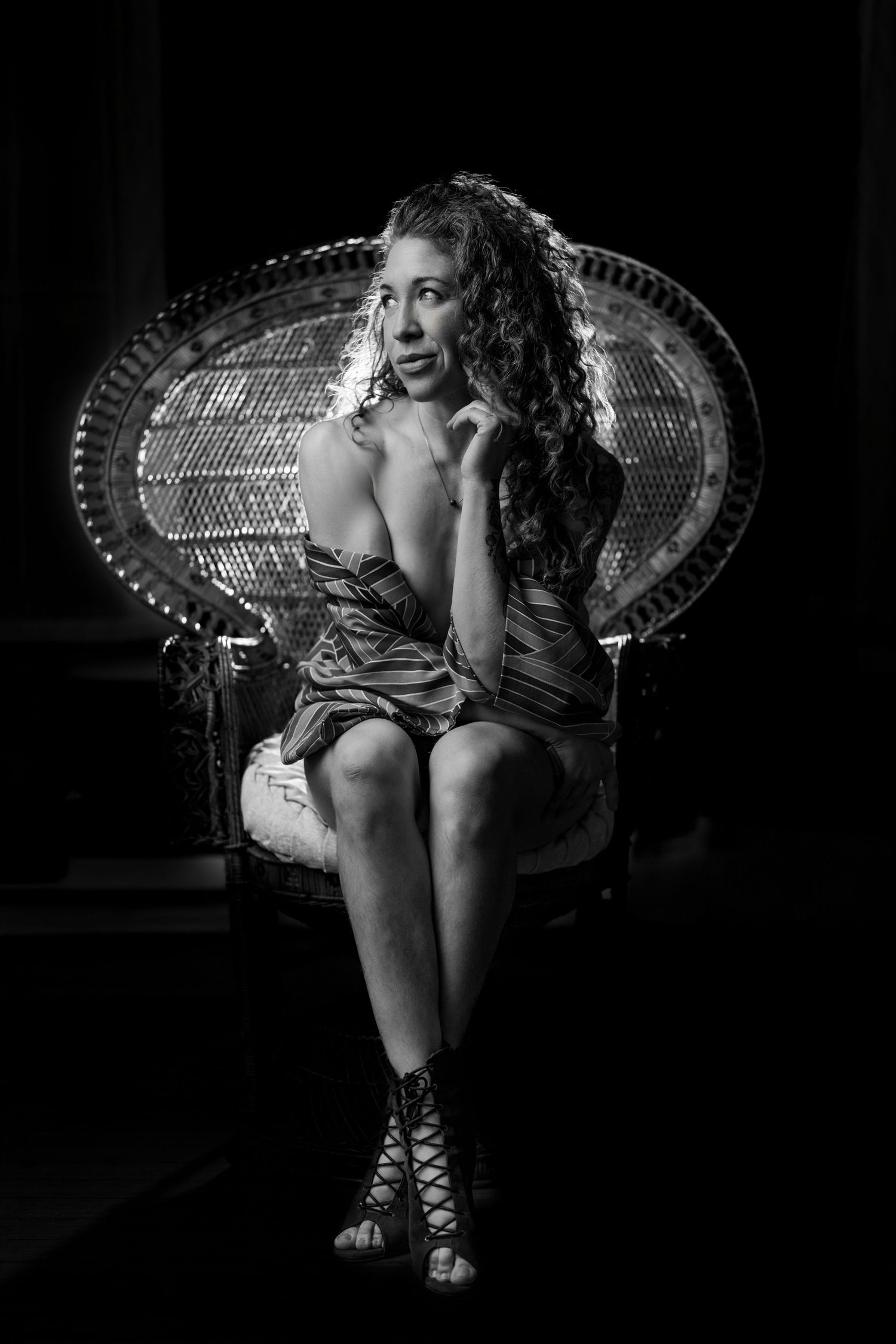 Black and white portrait of a curly-haired woman in a short dress sitting in a wicker peacock chair.