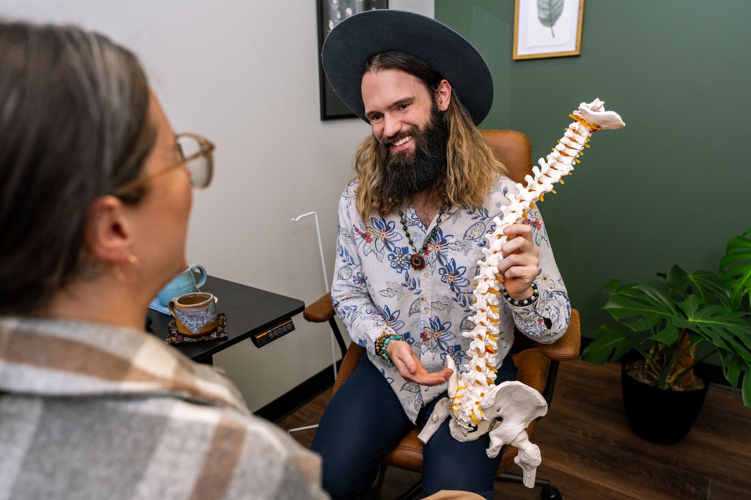 Bearded chiropractor in a hat shows a spinal model to a patient during a consultation in a modern clinic.