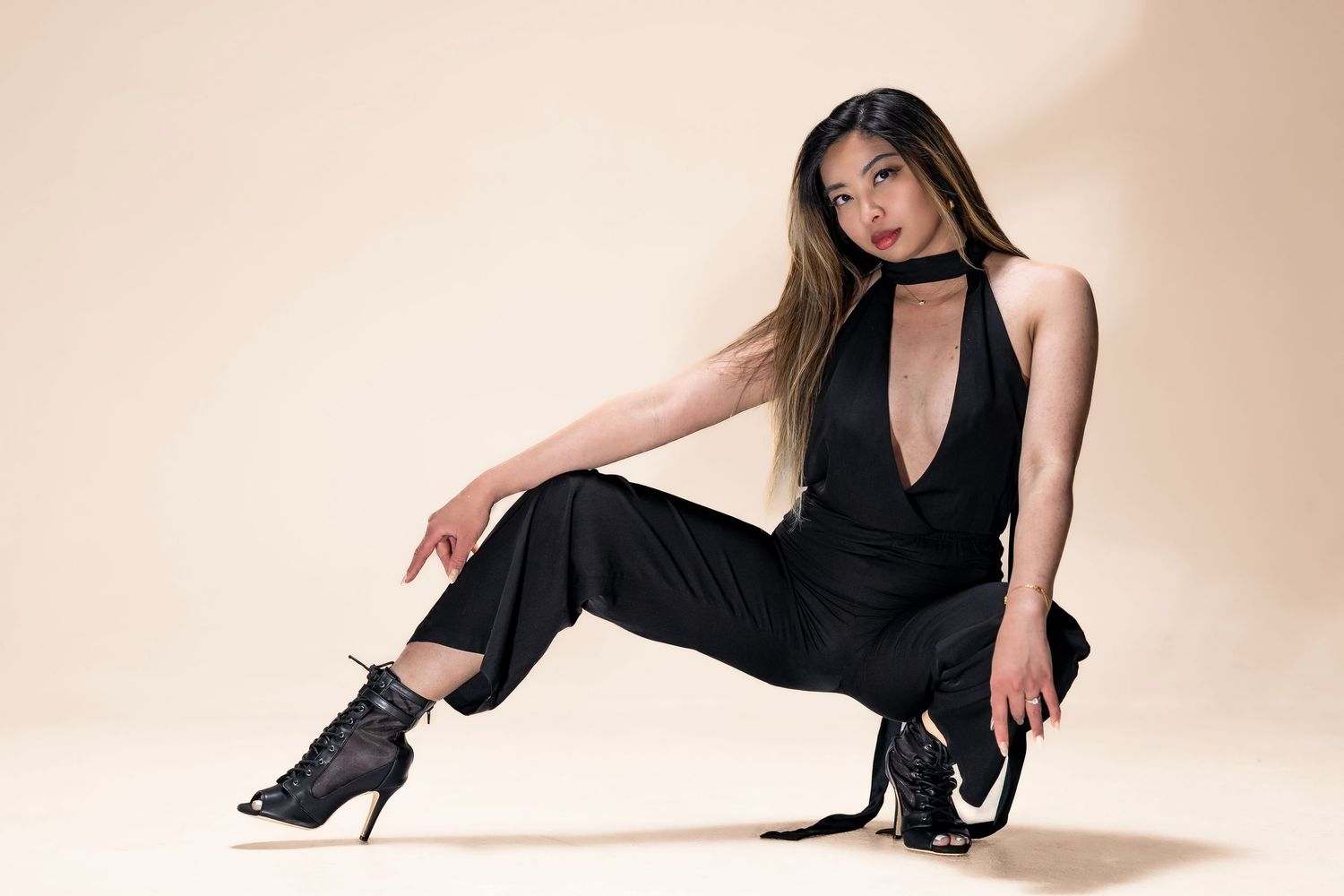 Asian woman in black plunging jumpsuit and heeled boots posing on studio floor.