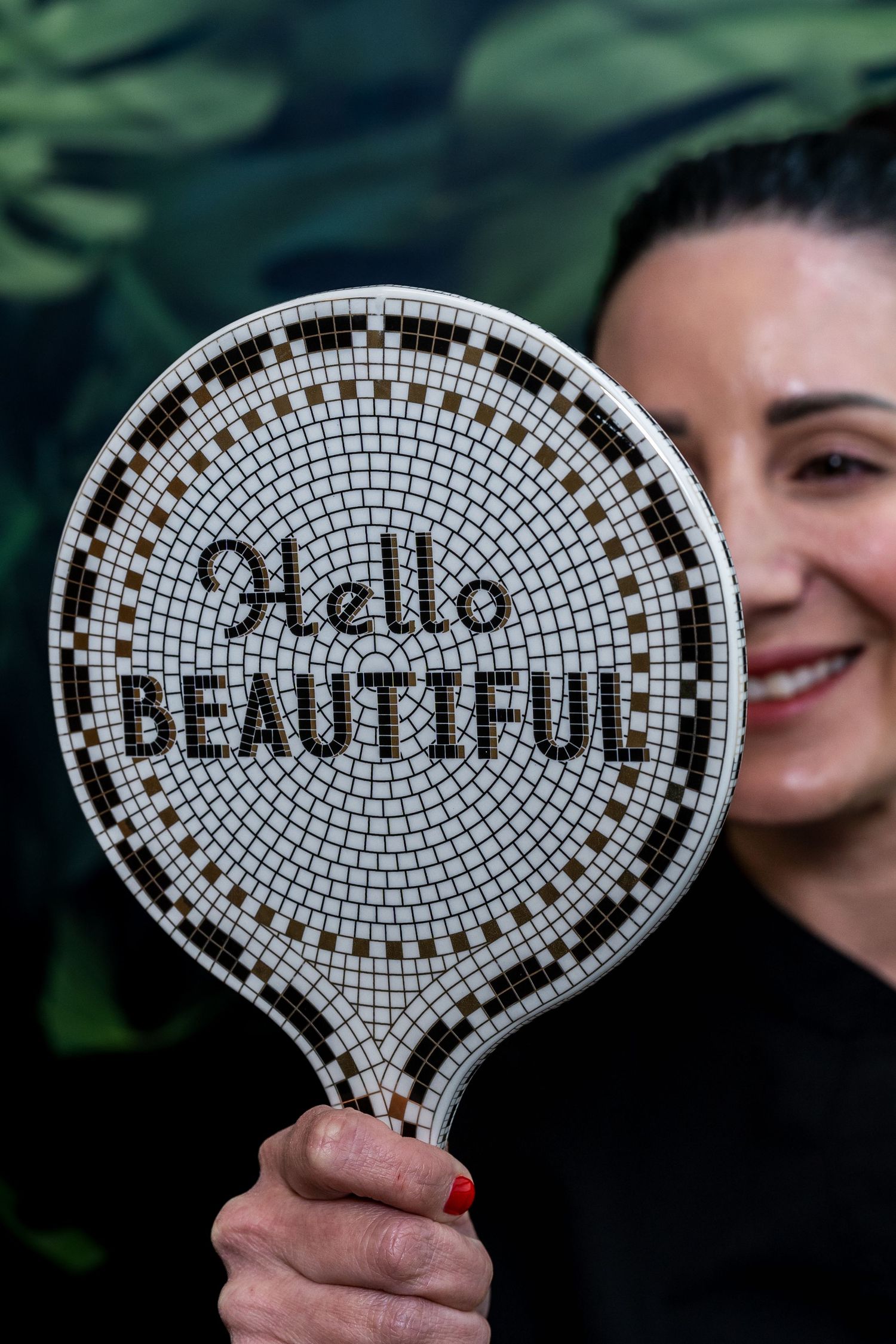 Smiling woman holding a mosaic tile hand mirror with Hello Beautiful text, green leafy background.