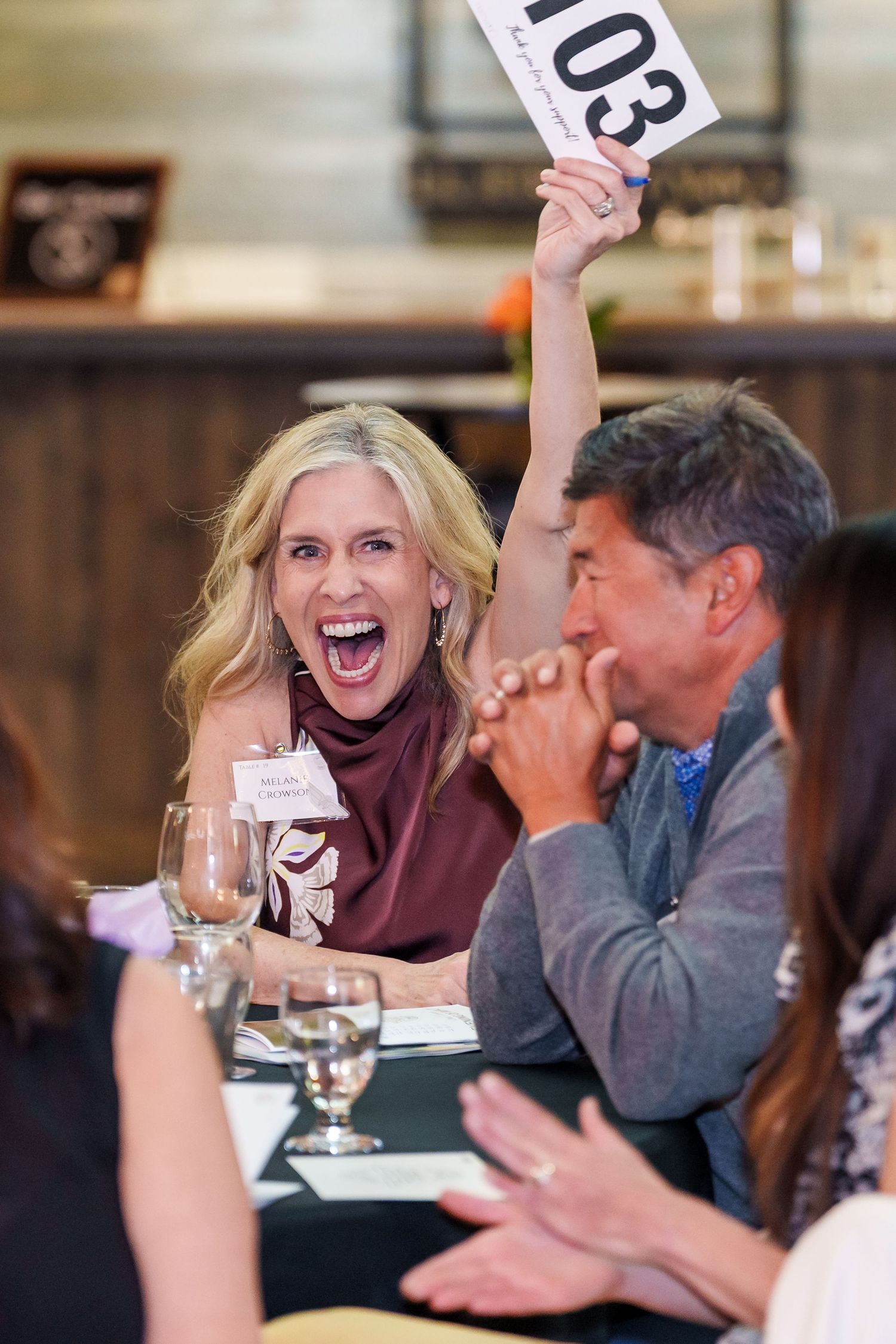 Excited blonde woman raises auction paddle 103 at charity event while man beside her laughs.