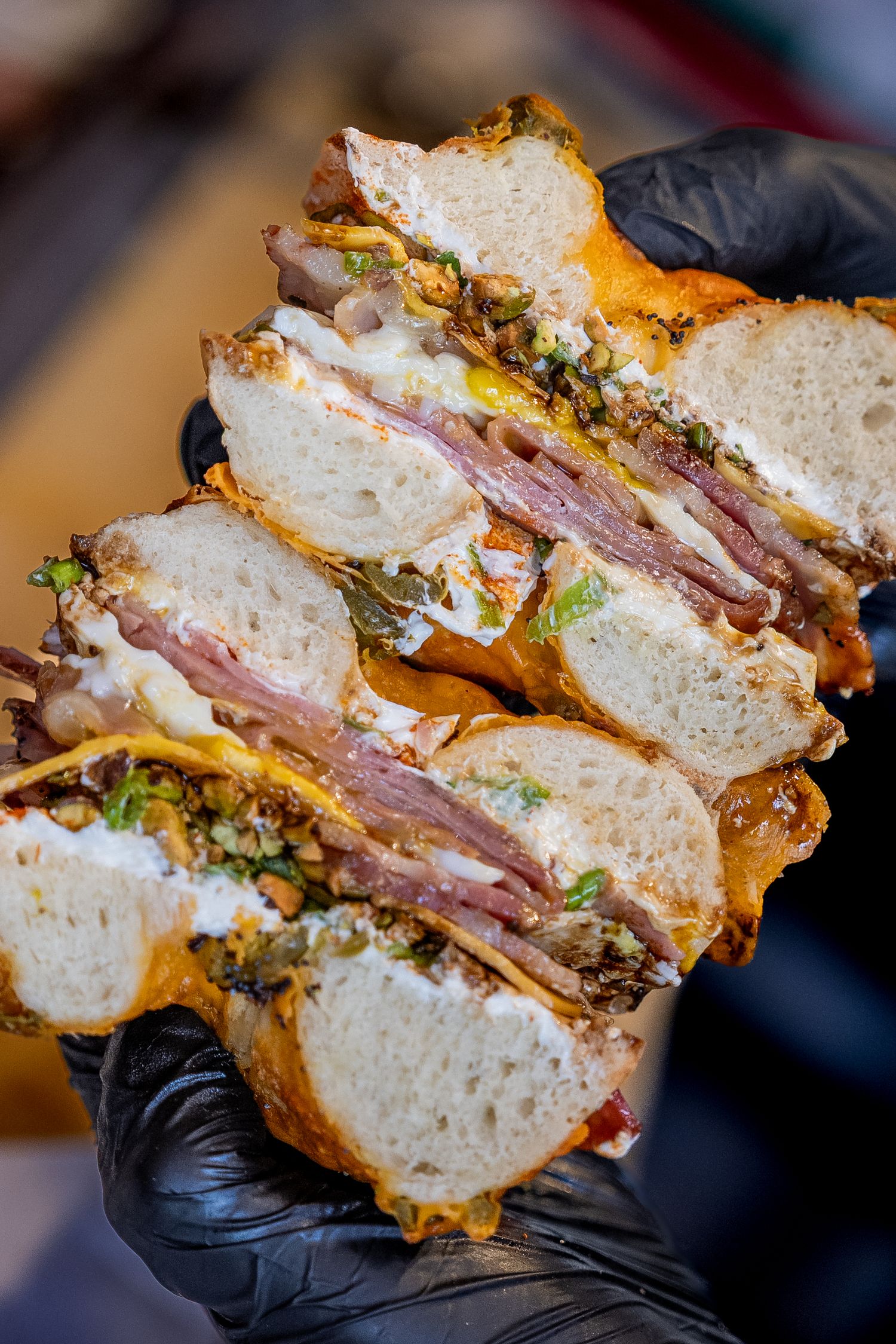 Gloved hands holding a thick, stacked sandwich with roast beef, melted cheese, and fresh toppings on toasted bread.