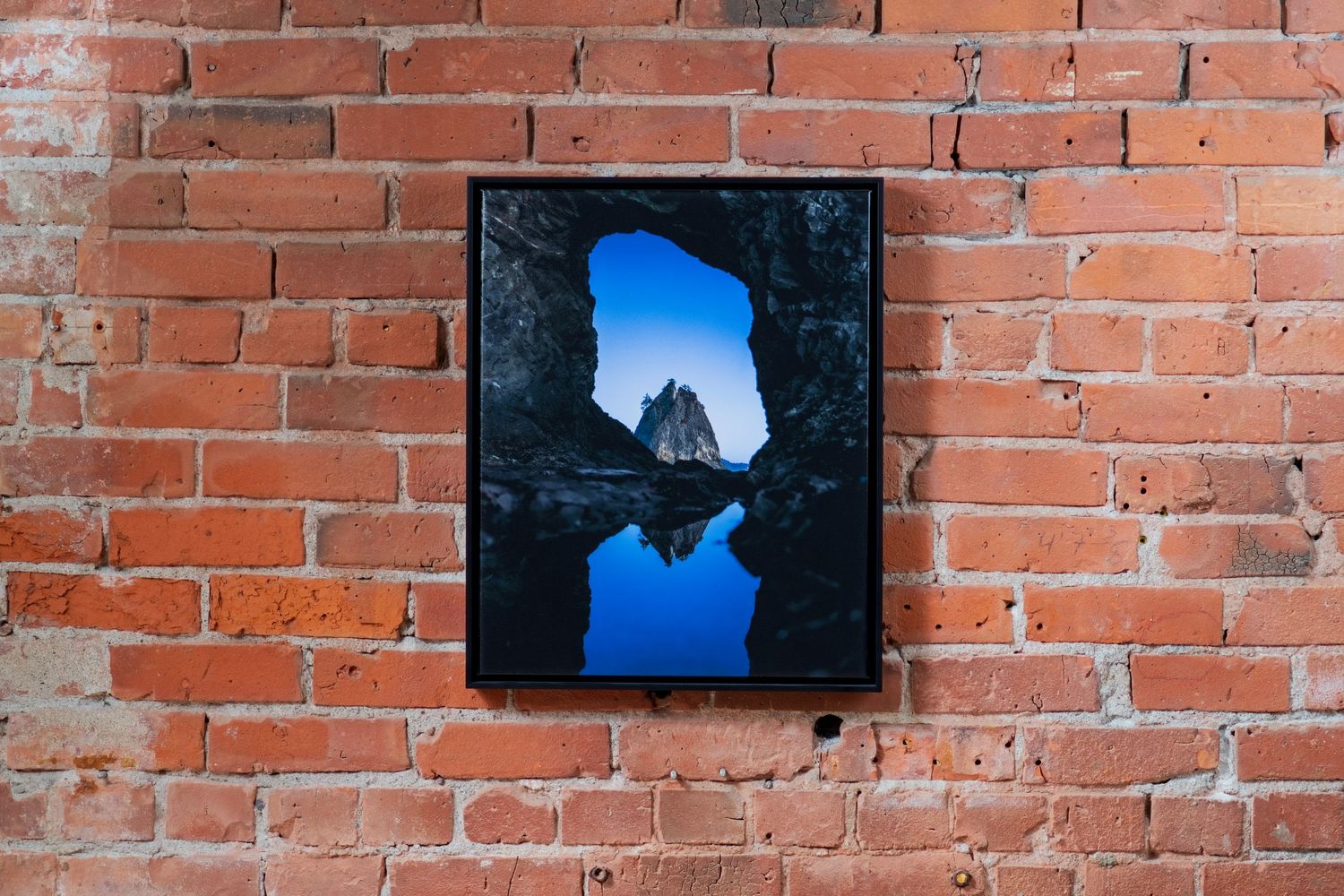 A framed photo of a rocky cave opening reflecting blue sky hangs on a rustic red brick wall.
