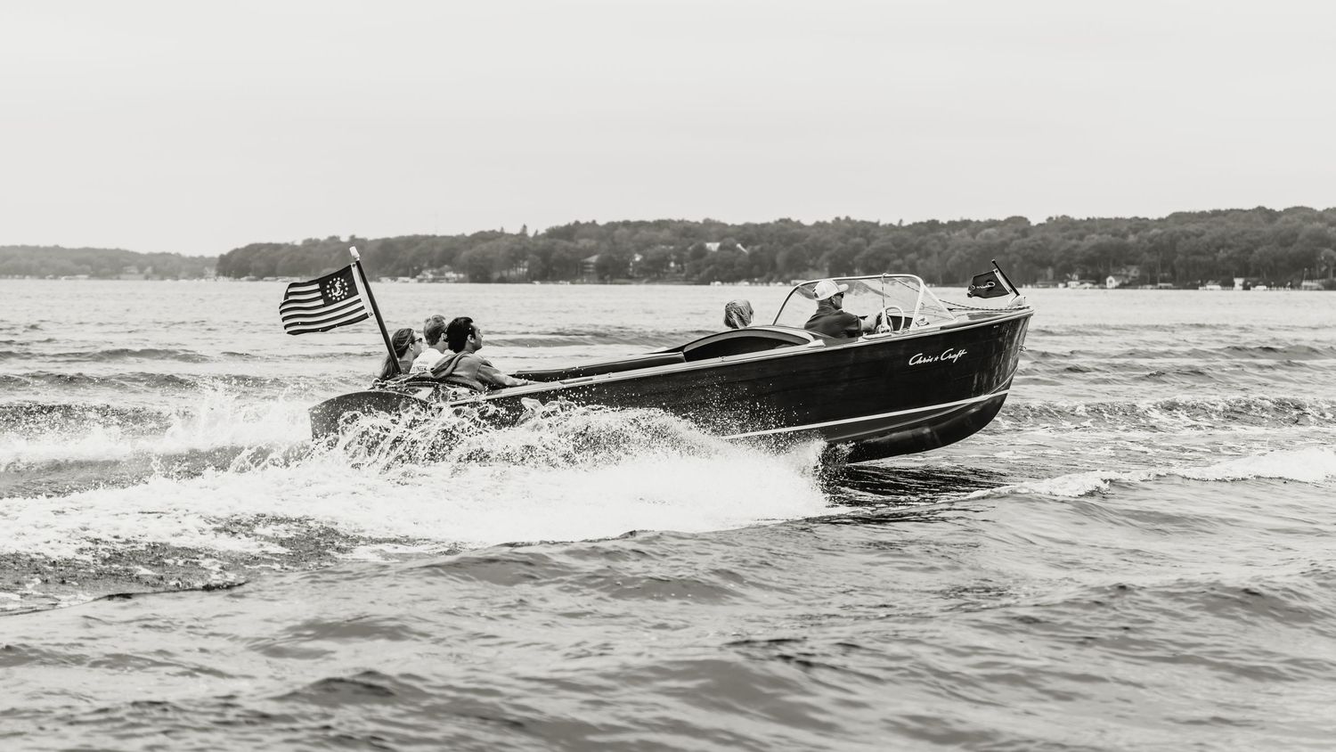 Classic wooden speedboat with American flag cuts through choppy water in vintage black and white photo.