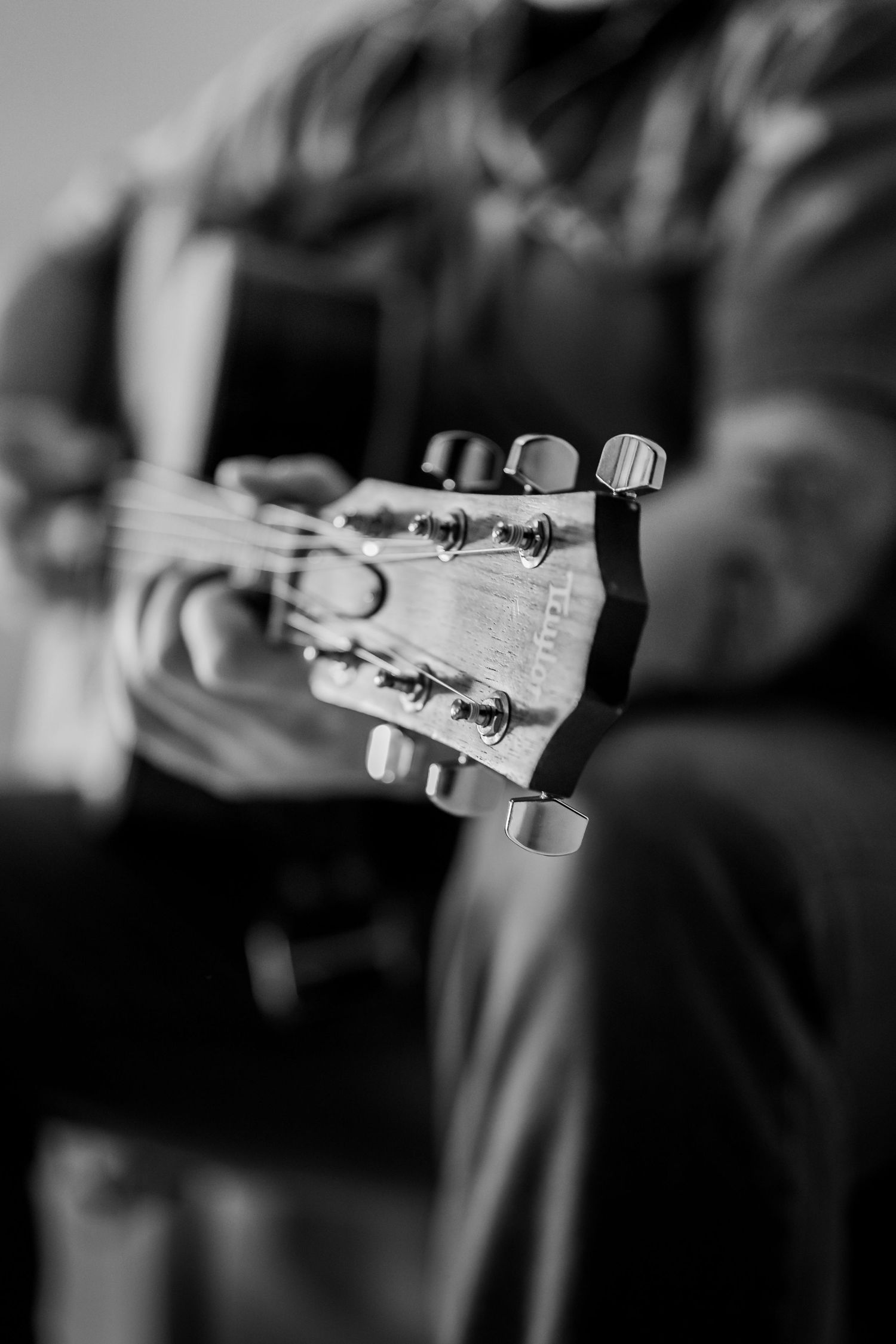 Black and white close-up of a guitarist playing acoustic guitar, focusing on the headstock and tuning pegs.