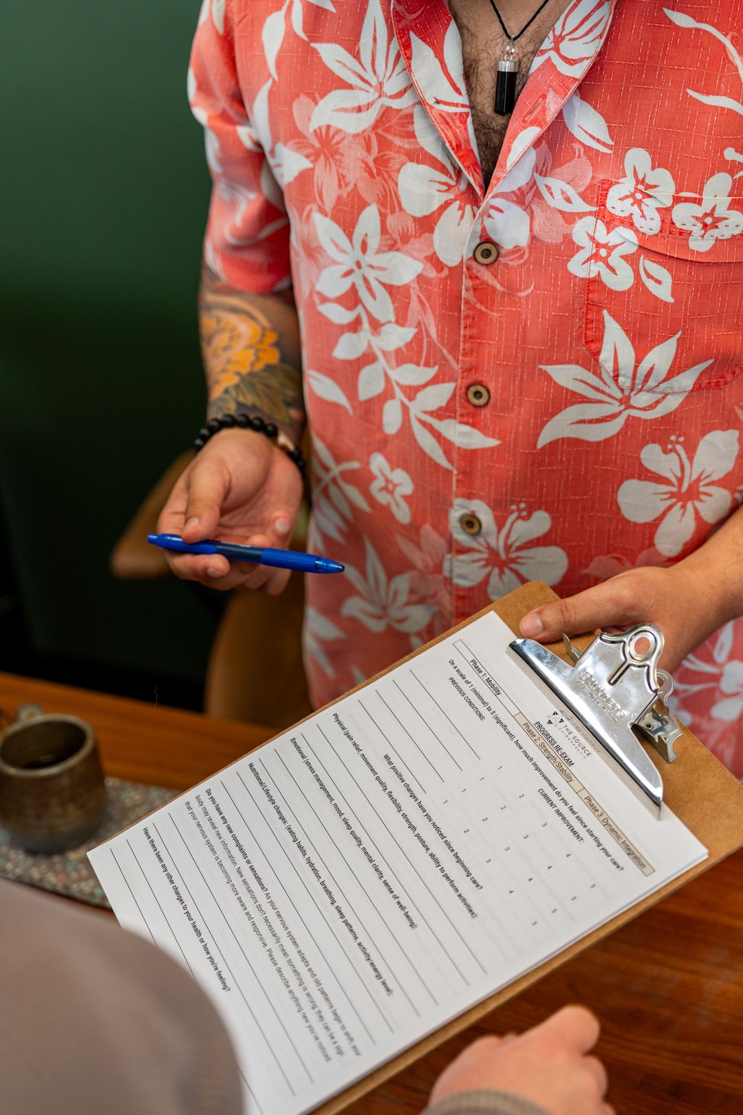 Person in floral Hawaiian shirt holding a pen over a clipboard with a form at a wooden desk.