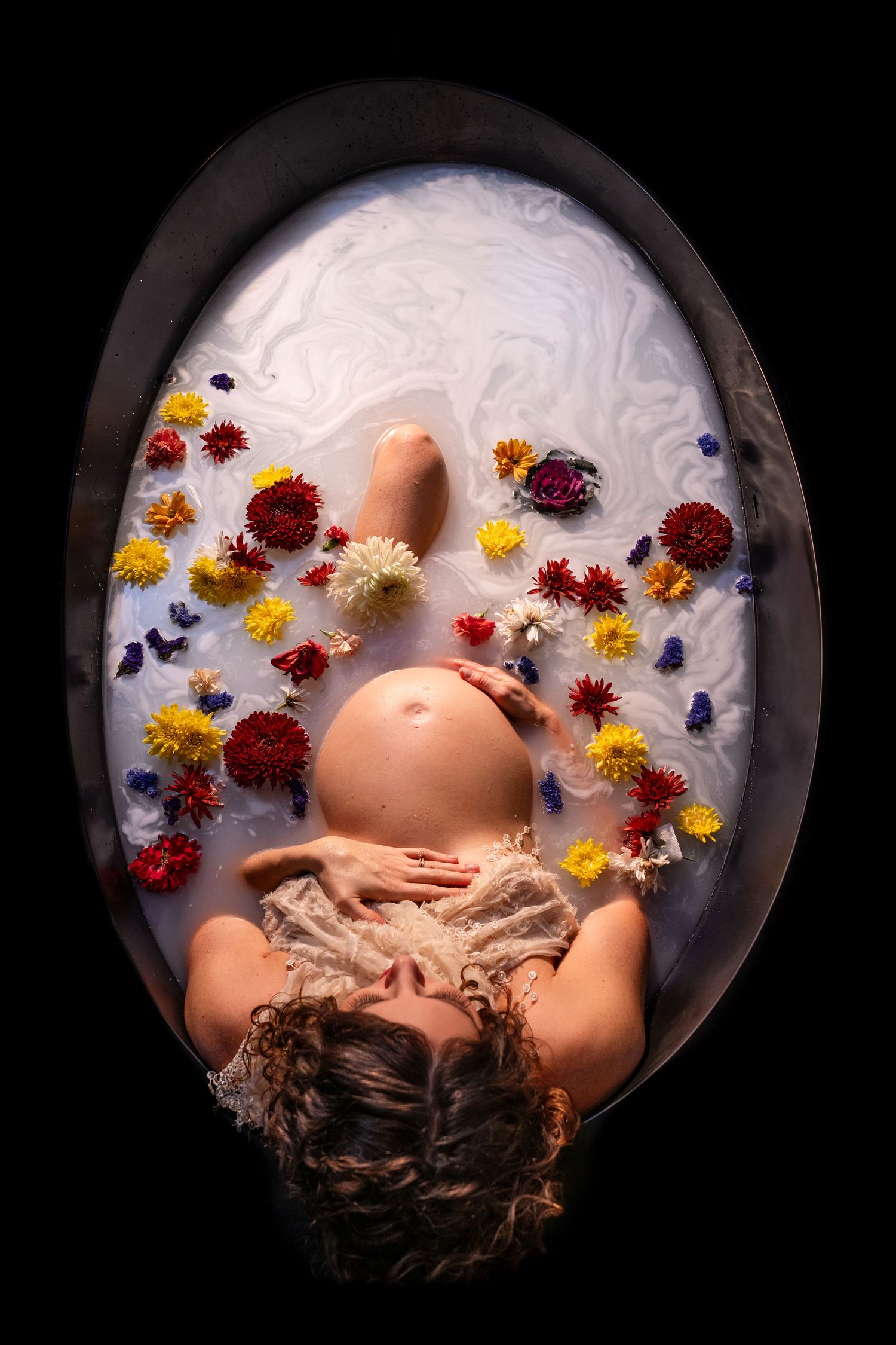 An overhead view of a pregnant woman relaxing in a milk bath surrounded by colorful flowers.