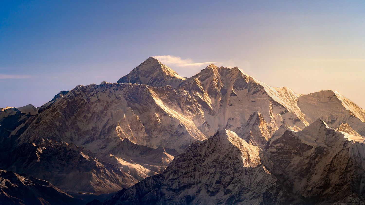 Majestic snow-capped mountain peaks bathed in golden sunset light against a clear blue sky.