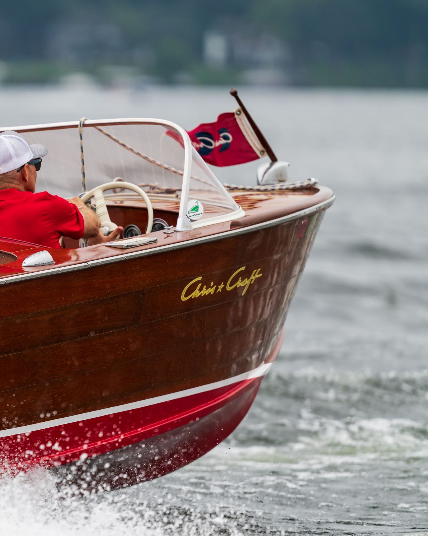 A classic Chris-Craft wooden speedboat with red hull navigates choppy waters, piloted by a man in a red shirt.