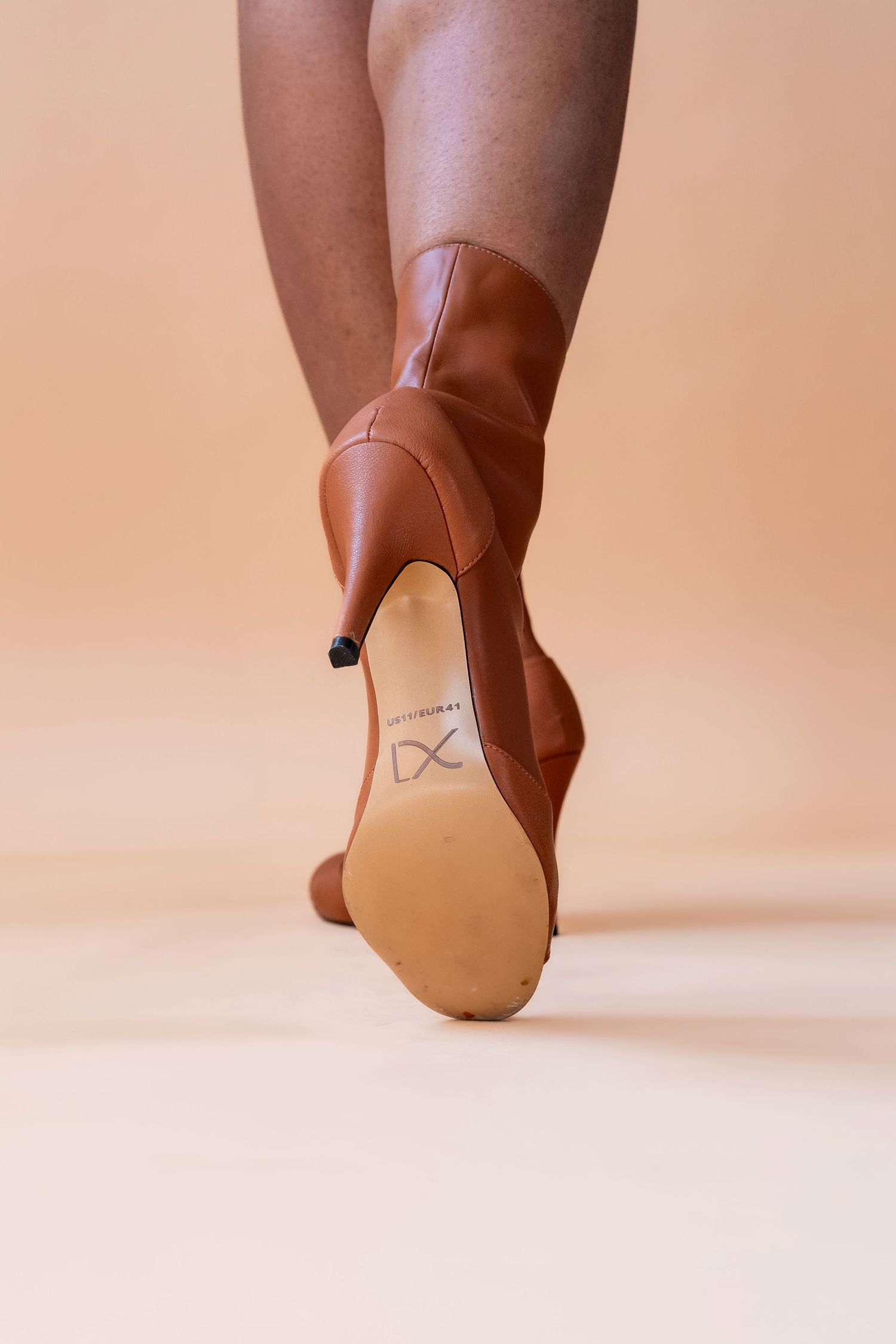 Brown leather stiletto boots shown from behind on a beige background, displaying the sole and heel detail.