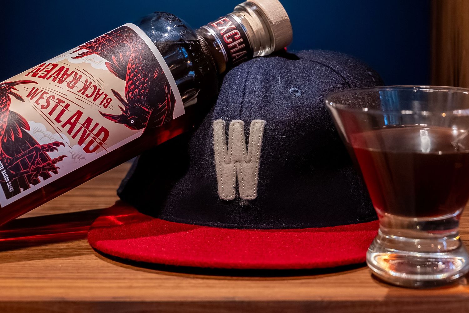 Westland Whiskey bottle beside a navy and red W snapback cap and a glass of whiskey on a wooden surface.