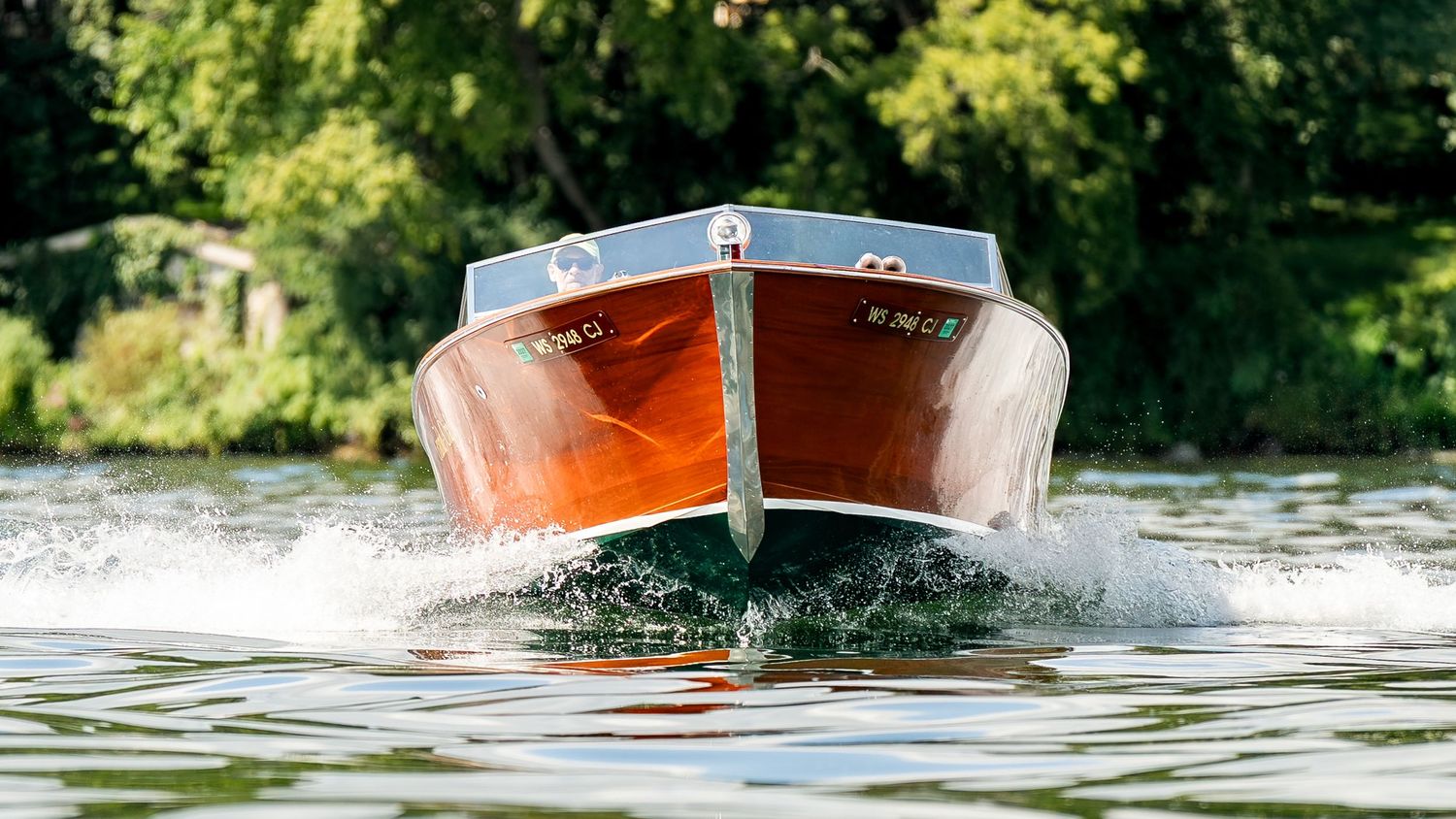 Classic wooden motorboat speeding across calm water with lush green trees in the background.