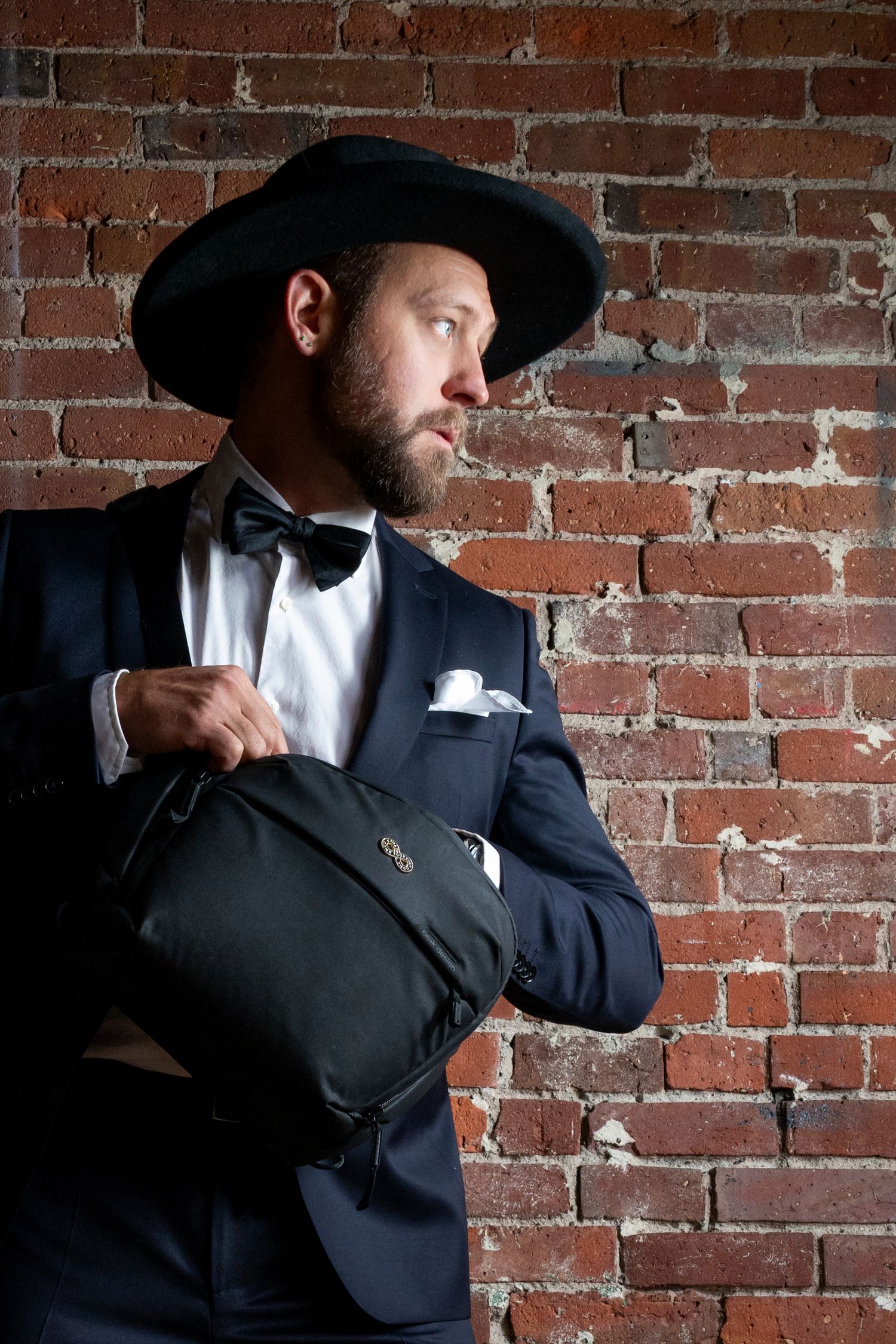 Bearded man in a navy suit, bow tie, and black hat carrying a bag against a brick wall.
