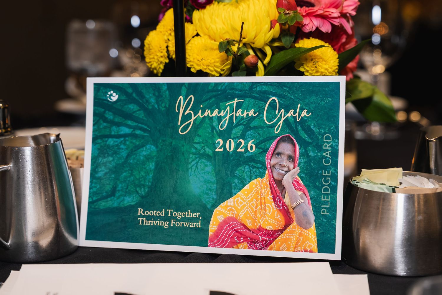 Binaystein Gala 2026 pledge card with teal design, reading Rooted Together, Thriving Forward, beside floral centerpiece.