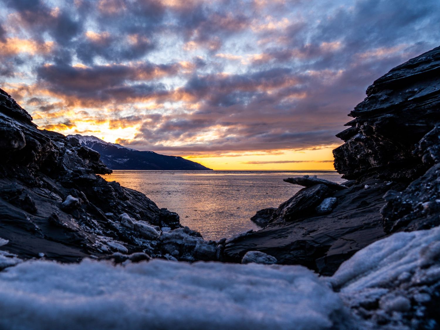 Dramatic Arctic sunset over rocky coastline with snow-dusted boulders framing a calm sea under vibrant cloudy skies.