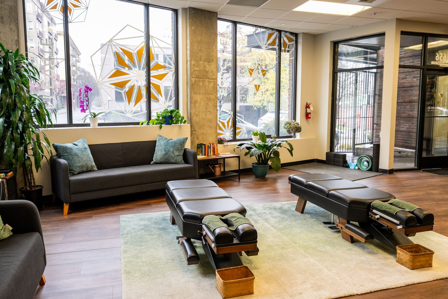Modern chiropractic office waiting room with gray sofas, adjustment tables, hardwood floors, and decorative floral window decals.