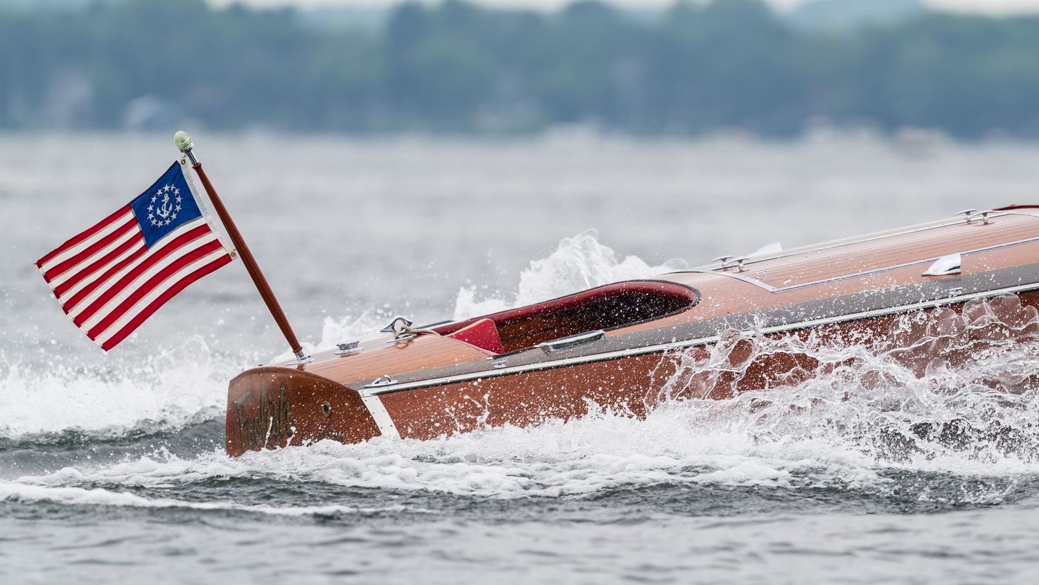 Classic wooden speedboat racing across choppy water with an American flag flying from the stern.