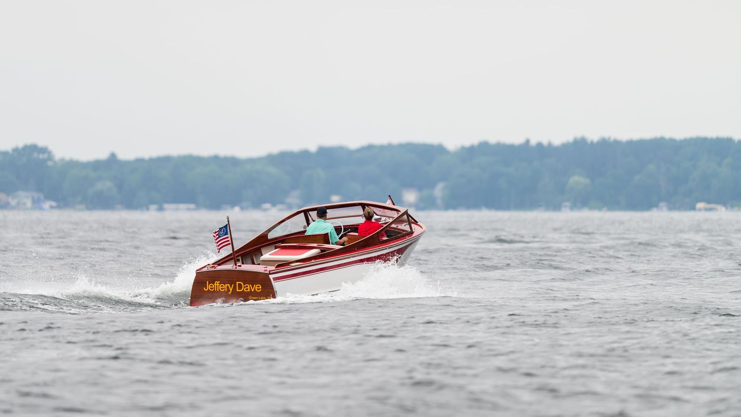 Classic red wooden speedboat named Jeffrey Clever cruising on calm lake waters with American flag flying.