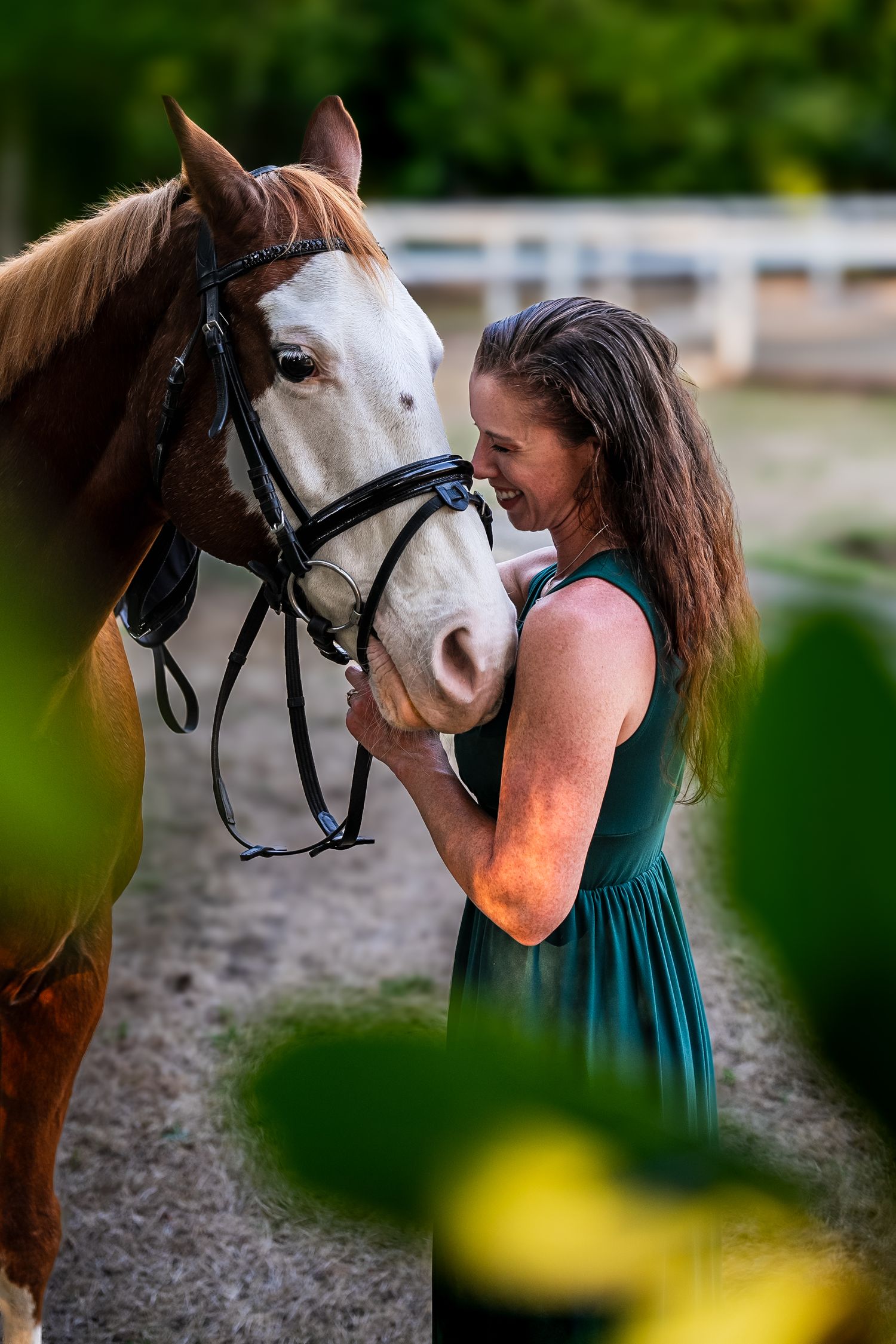 Woman in green dress smiling warmly while holding the bridle of a chestnut horse outdoors.