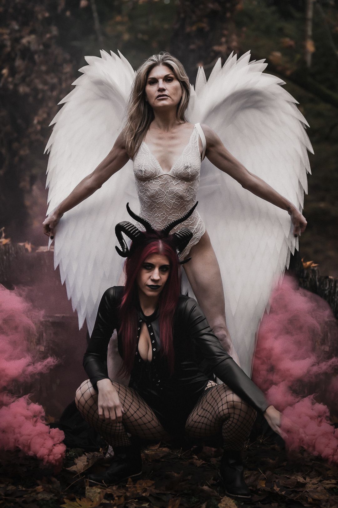 Two women cosplay as an angel with white wings and a devil with horns amid pink smoke in a dark forest.