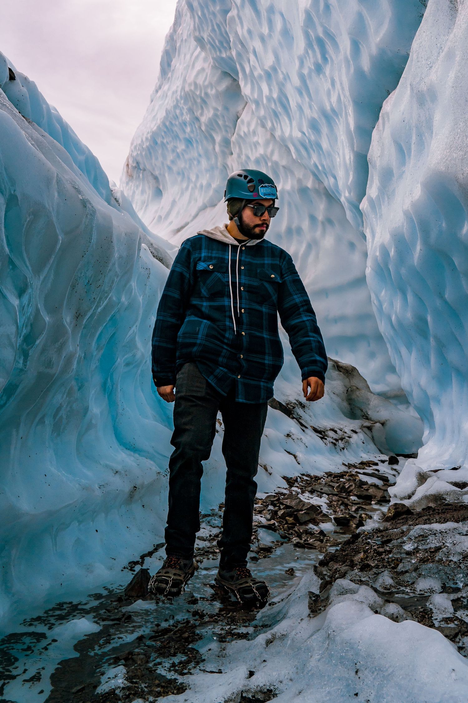 A hiker in a plaid jacket and helmet walks through a narrow glacial ice canyon with towering blue ice walls.