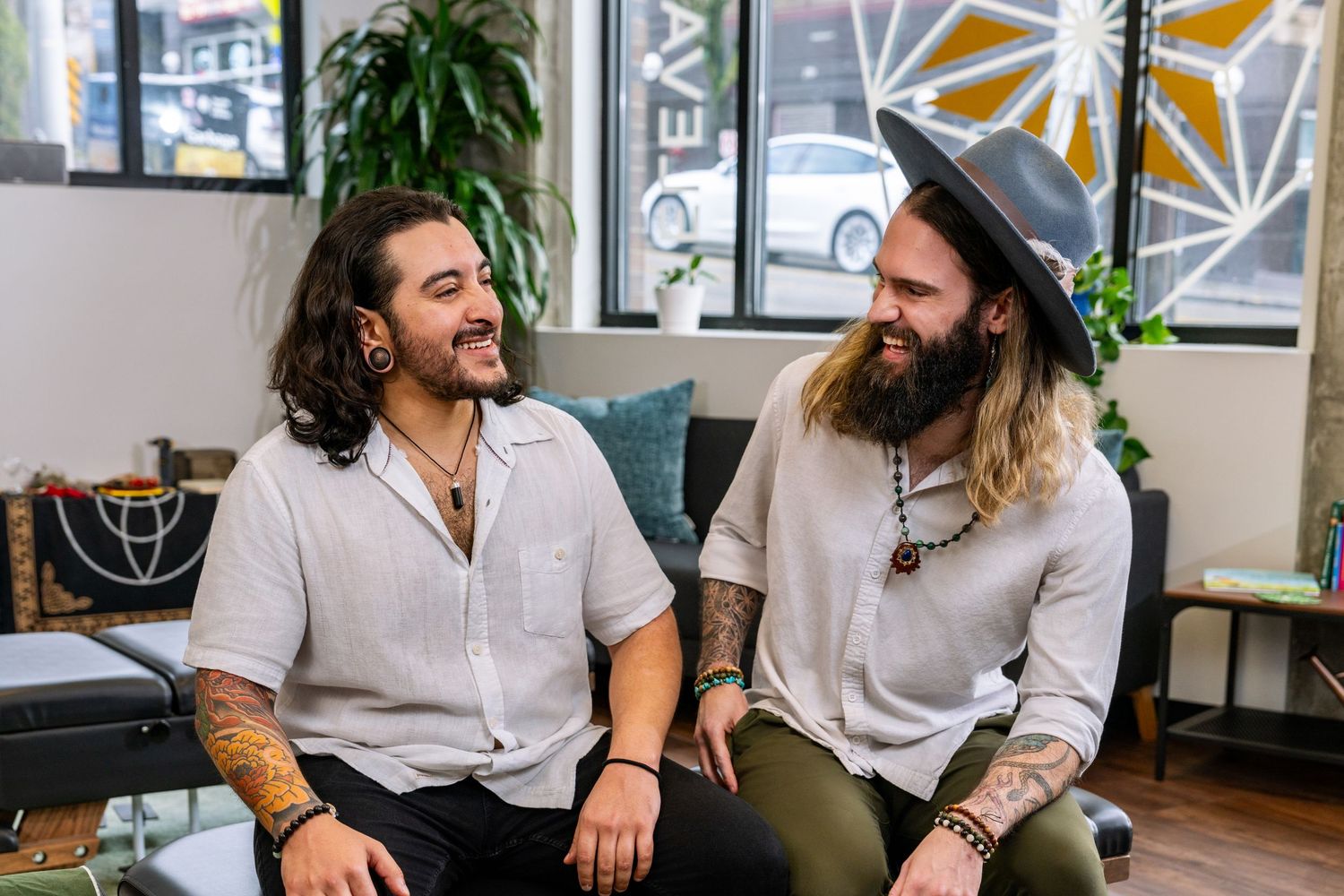Two tattooed men with long hair smiling and conversing in a modern wellness studio waiting area.