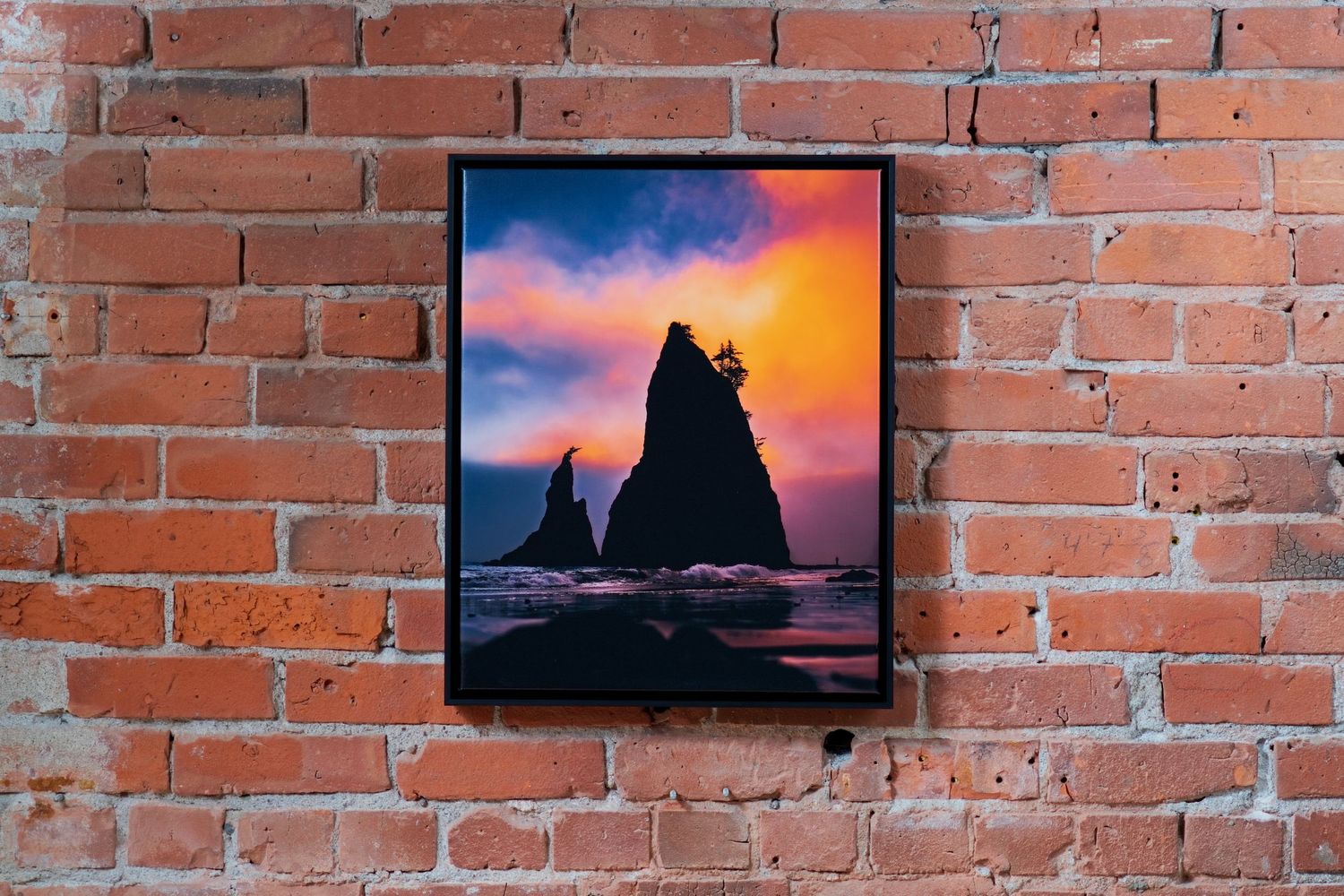 Framed coastal sunset photo with dramatic sea stacks silhouetted against vibrant purple and orange sky, displayed on brick wall.