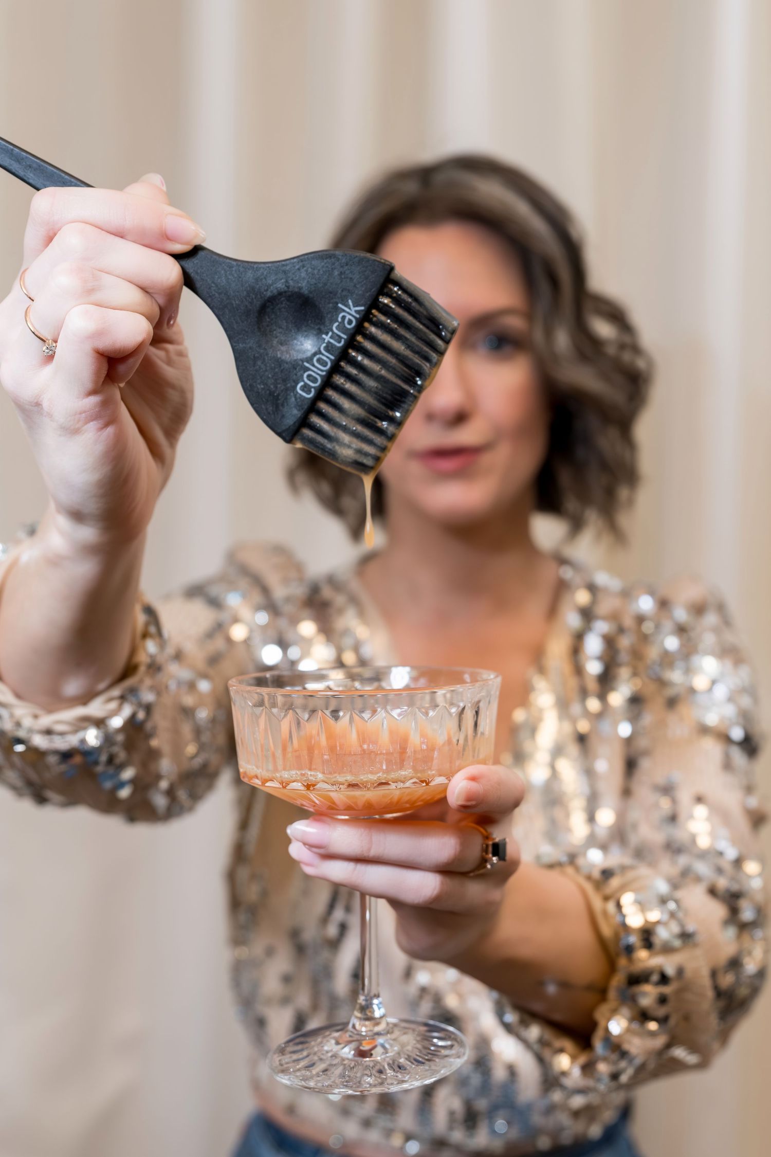 Woman in sequin top holding a cocktail glass while dipping a hair color brush into the drink.