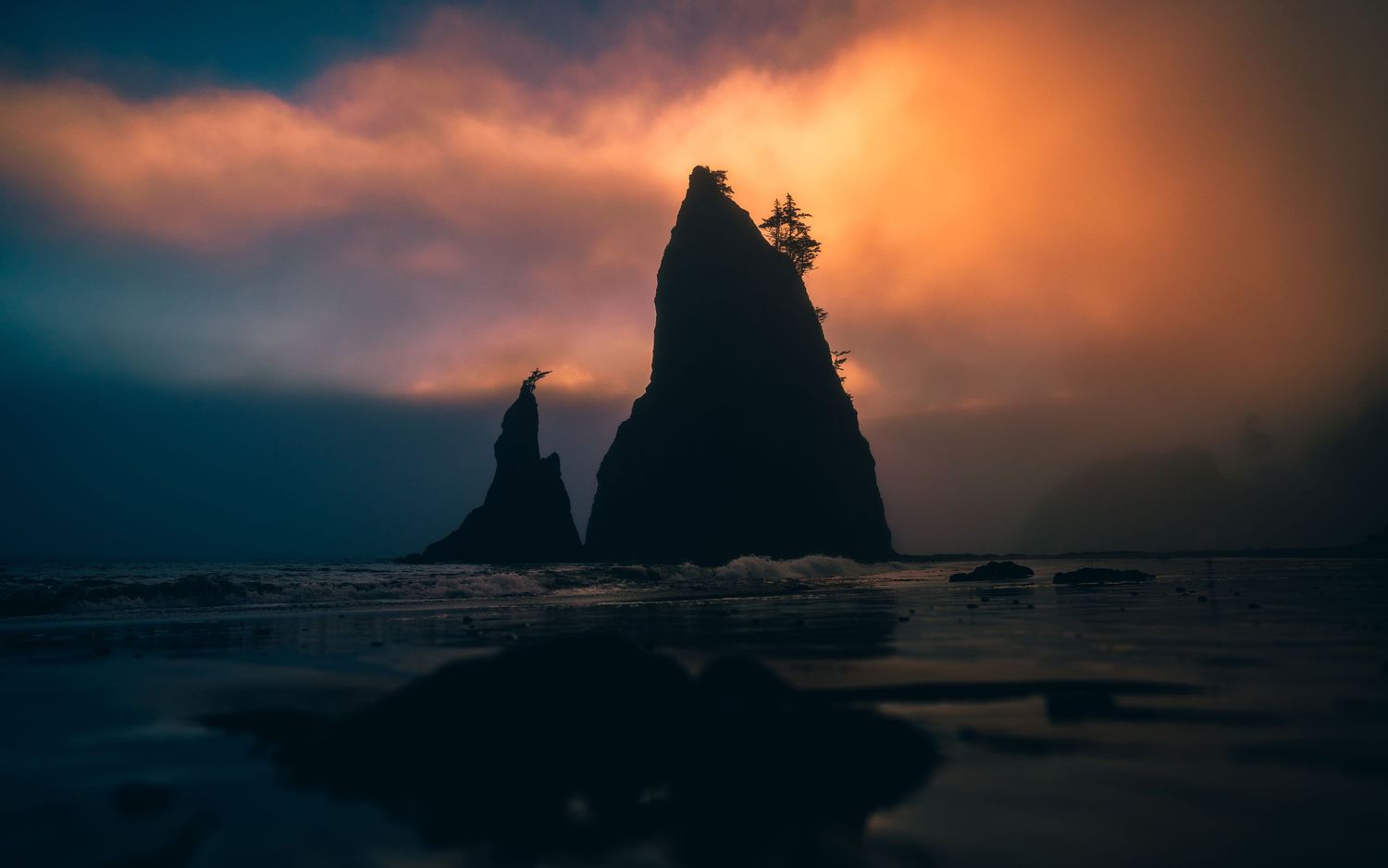 Dramatic sea stacks silhouetted against a fiery orange sunset sky with moody clouds over a dark ocean beach.
