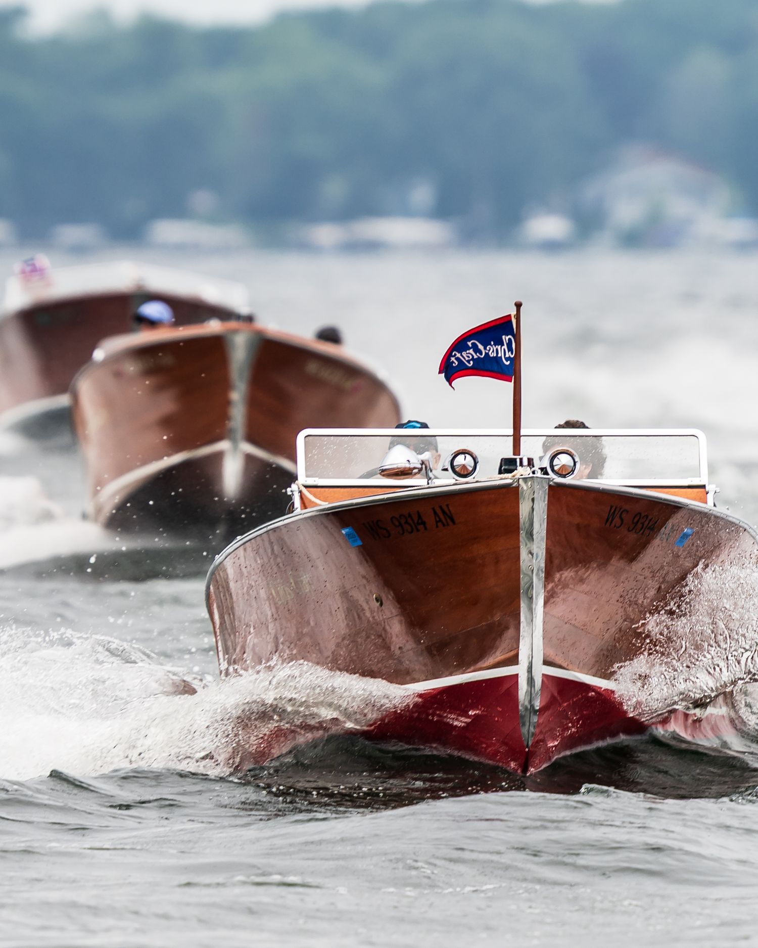 Vintage mahogany wooden classic boats race across a lake, led by a boat flying a red and blue flag.