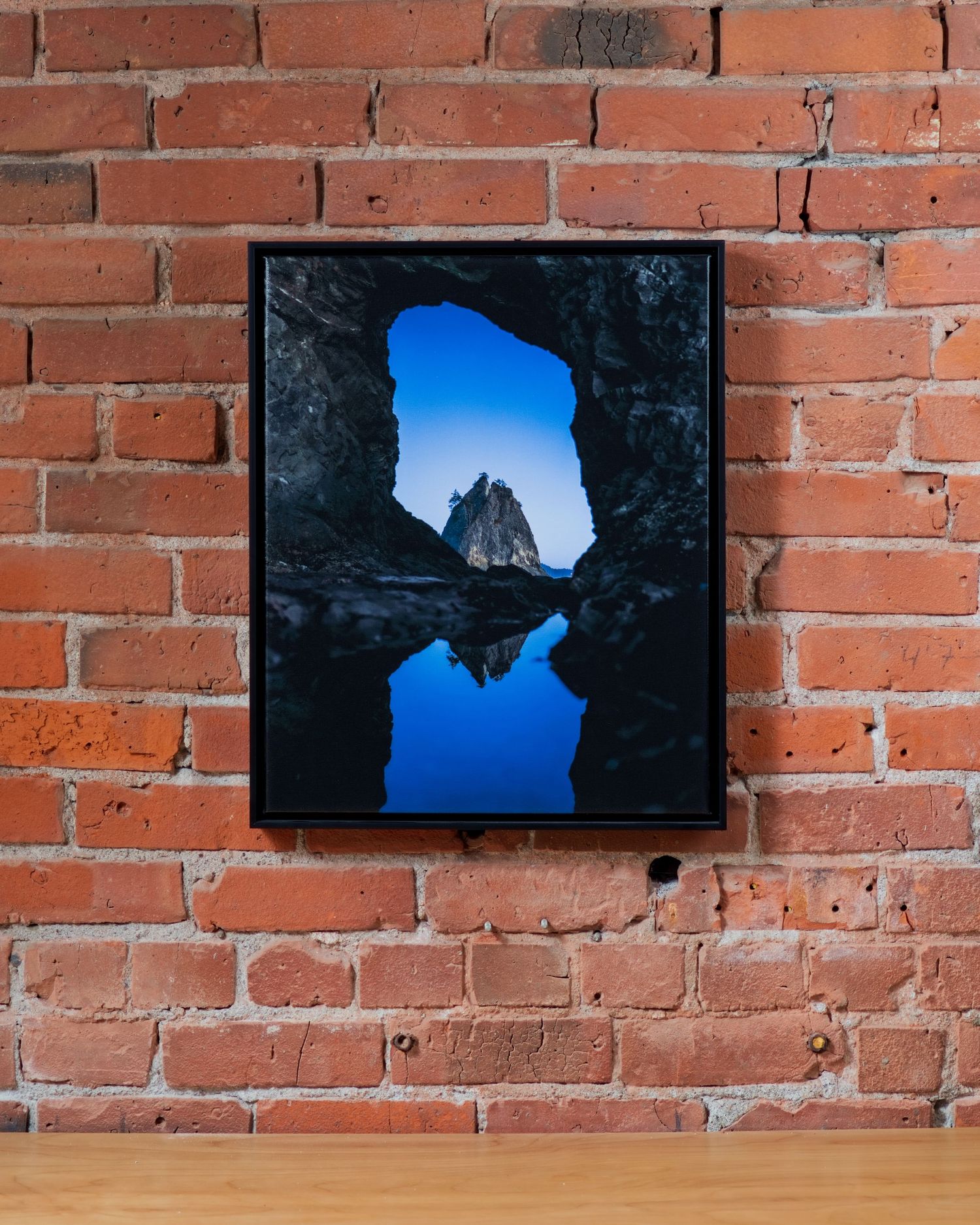 Framed photo of a rock arch reflecting in still water under a blue sky, displayed on a rustic brick wall.