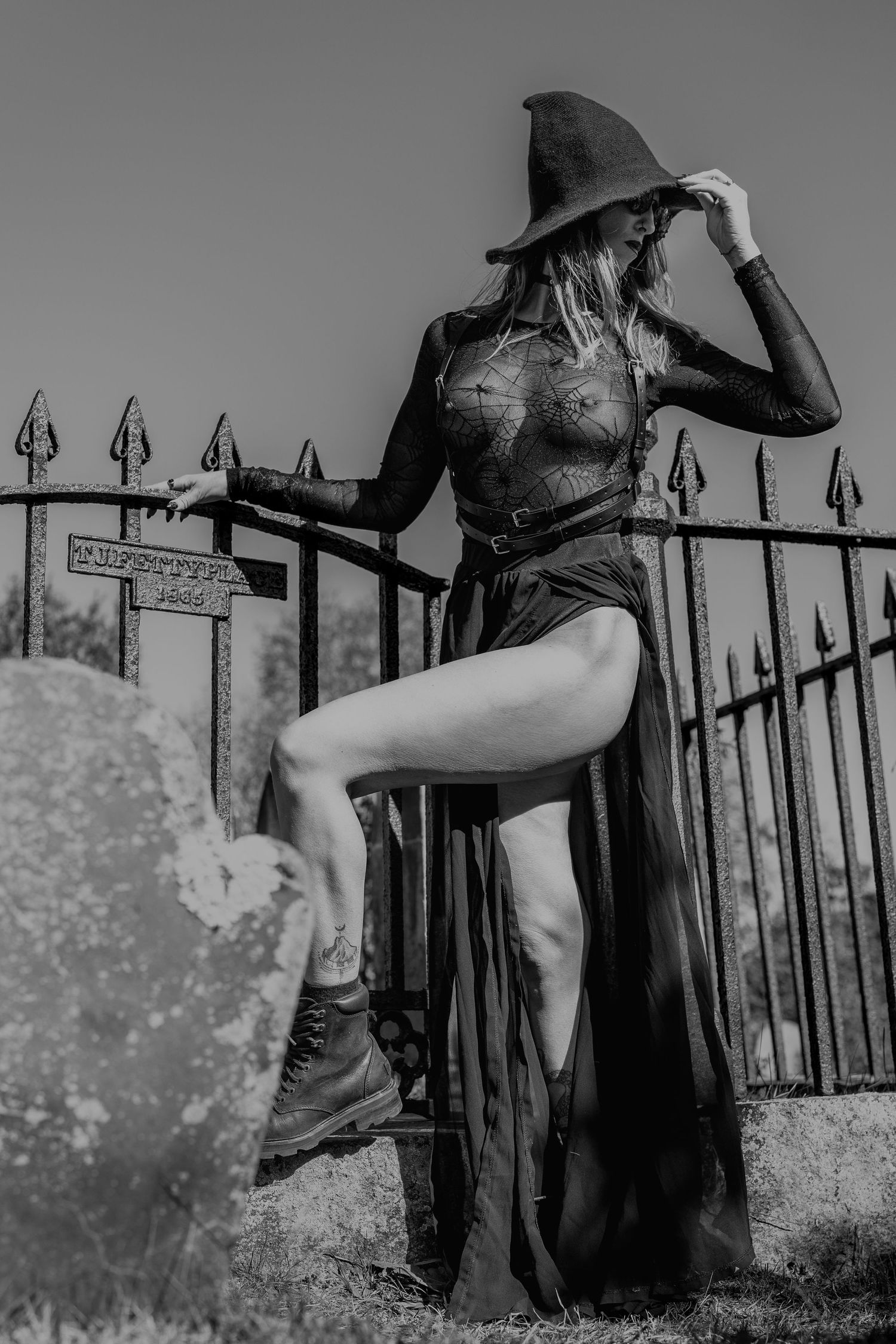 Black and white photo of woman in witch hat and sheer bodysuit posing at cemetery iron fence.