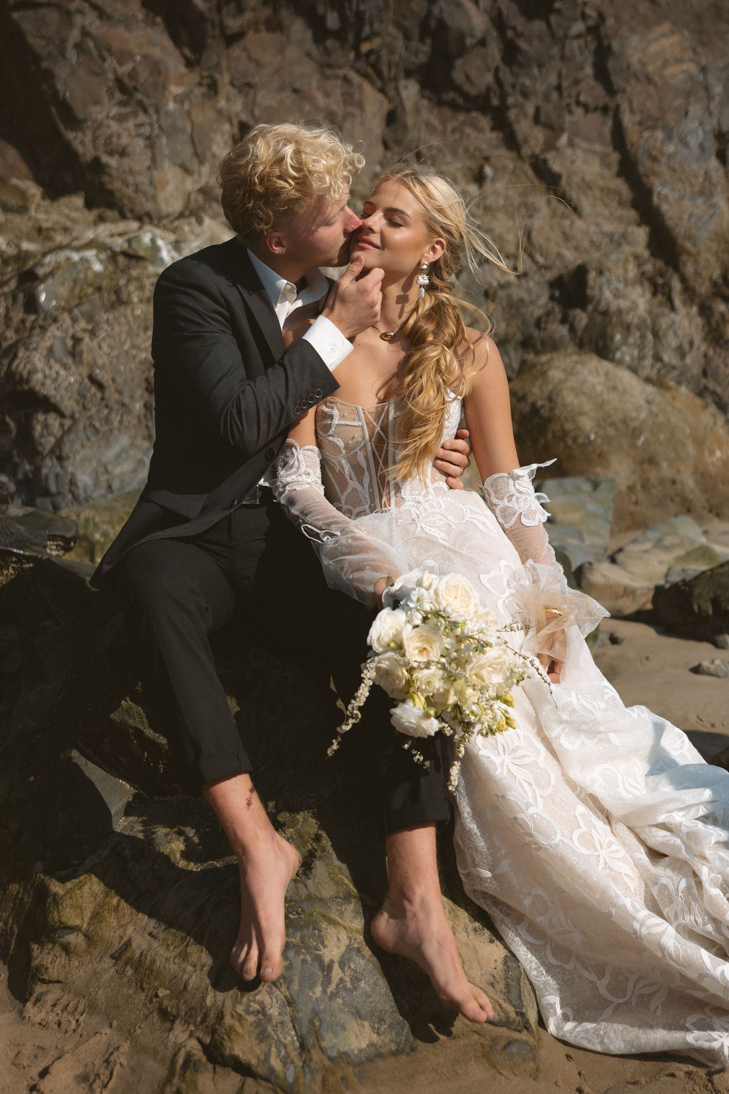 Newlyweds sharing romantic moments on rocky beach with white bouquet.