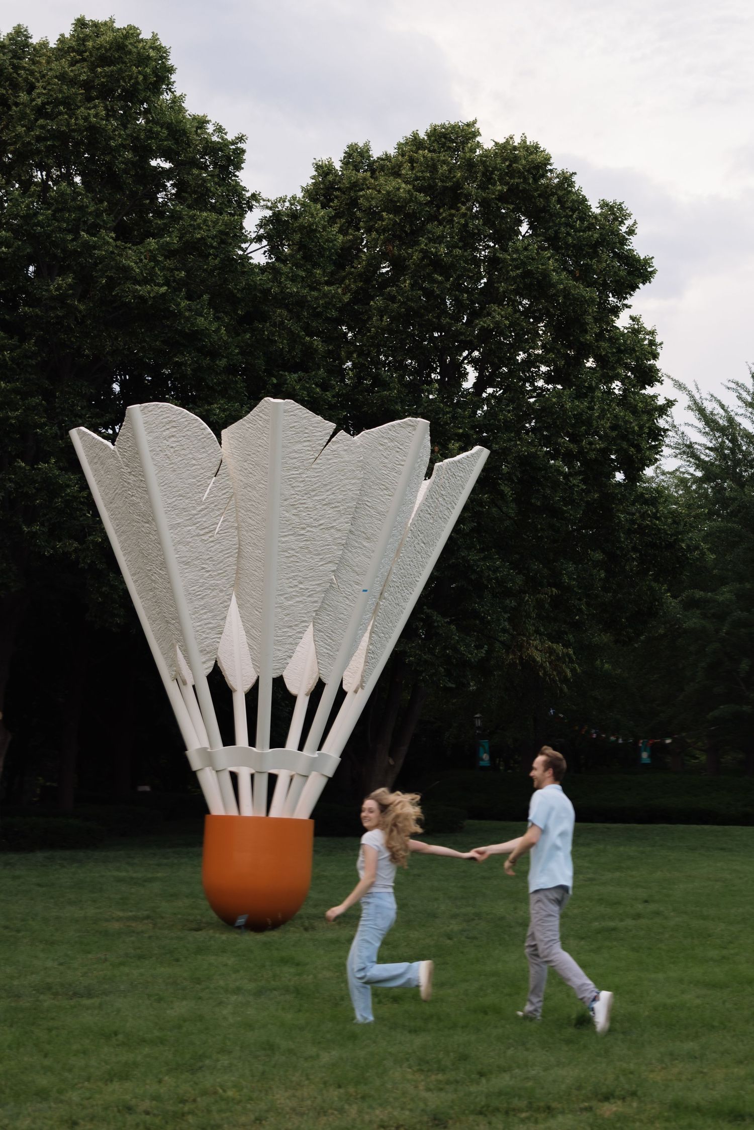 Couple holding hands running past a giant badminton shuttlecock sculpture in a green park.