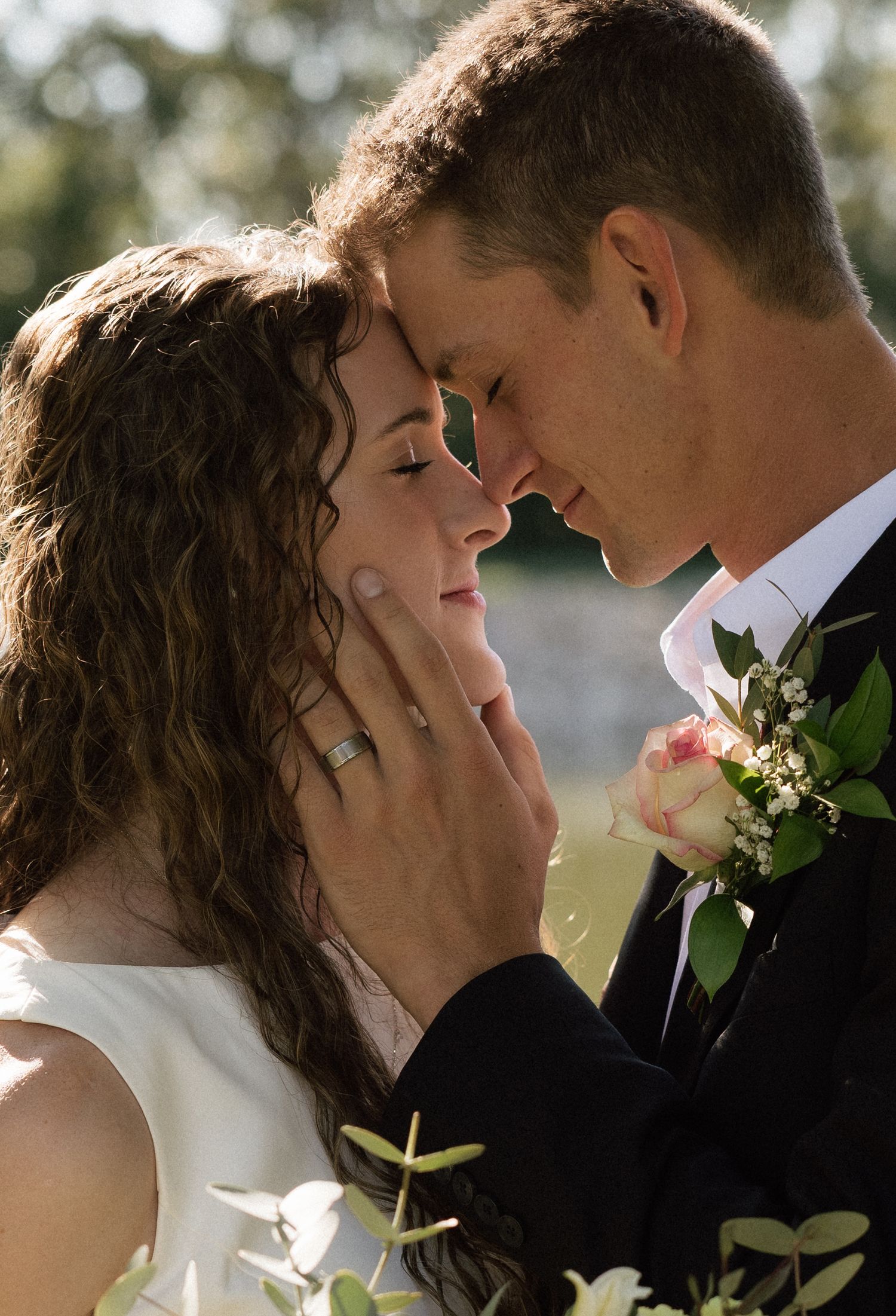 A groom tenderly cups his bride's face as they share an intimate moment, both wearing wedding rings, with floral boutonniere.