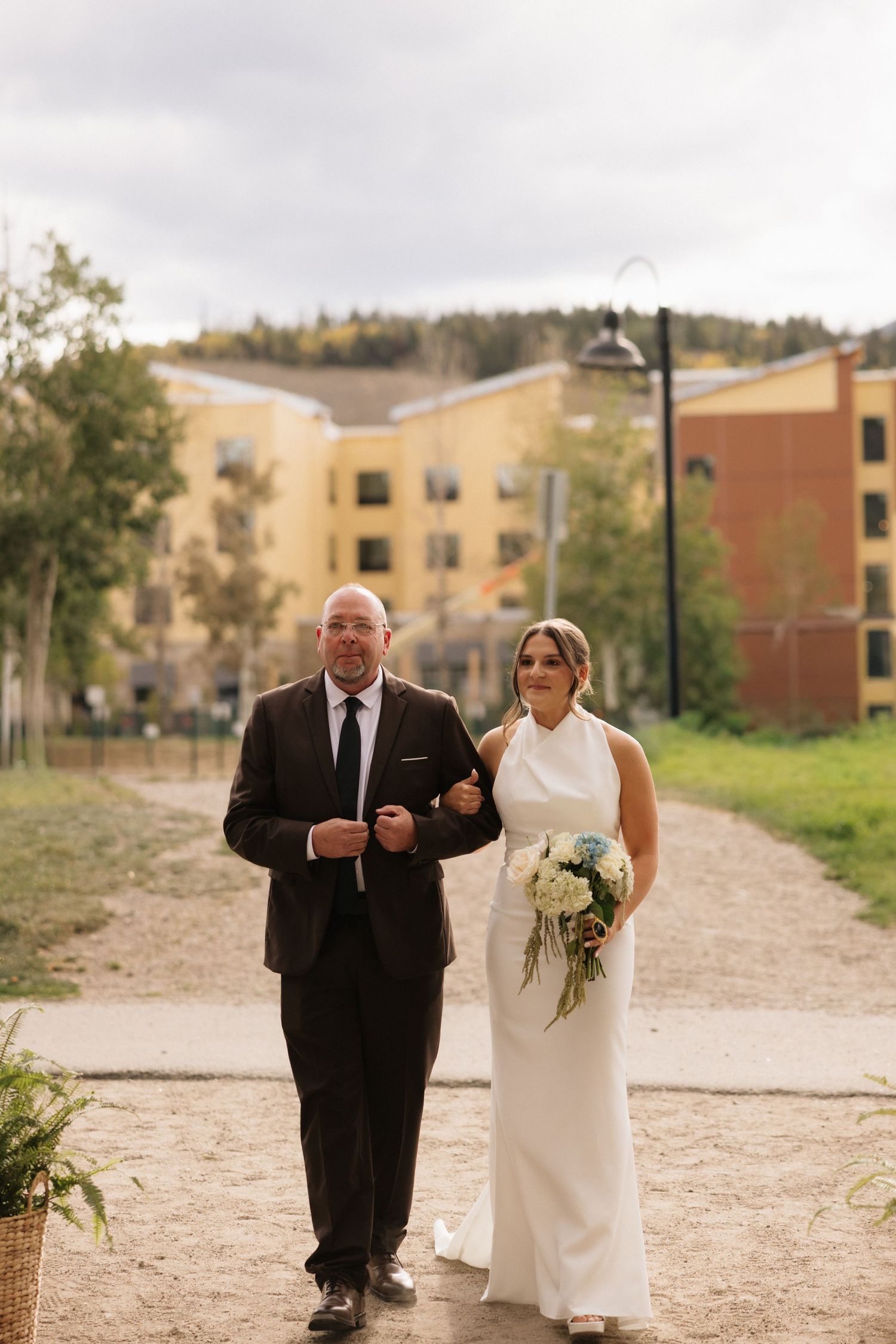 Bride in white halter gown holding bouquet walks with suited man on gravel path with buildings and trees behind them.