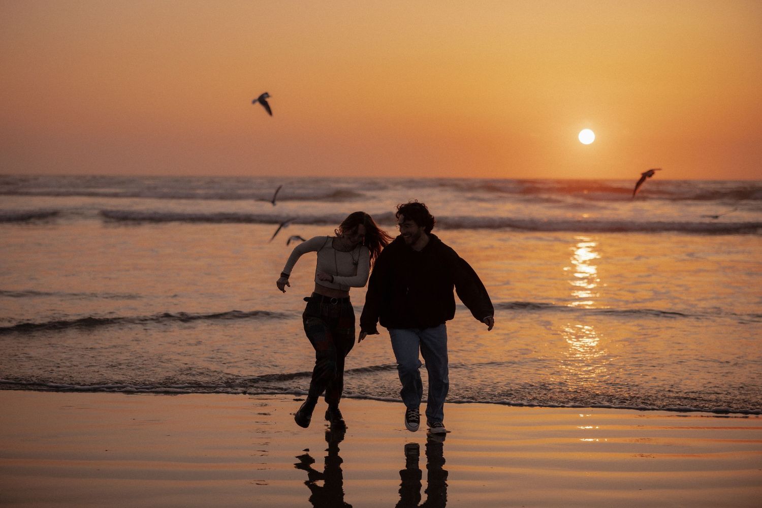 Silhouettes of people walking along a beach at sunset with seabirds flying overhead.